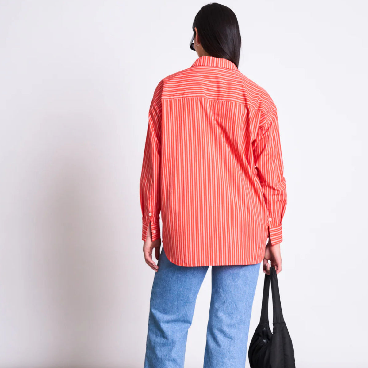 Person mit langen dunklen Haaren, die ein lockeres rot-weiß gestreiftes Hemd und blaue Jeans trägt und mit dem Rücken zur Kamera steht, während sie eine schwarze Tasche in der linken Hand hält, vor einem schlichten hellen Hintergrund.