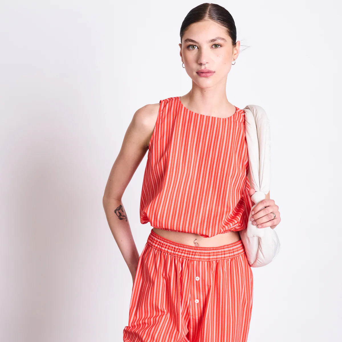 Eine Person, die ein ärmelloses, rot-weiß gestreiftes Crop-Top und eine dazu passende Hose trägt, steht vor einem unifarbenen Hintergrund und hält eine weiße Stepptasche über einer Schulter.