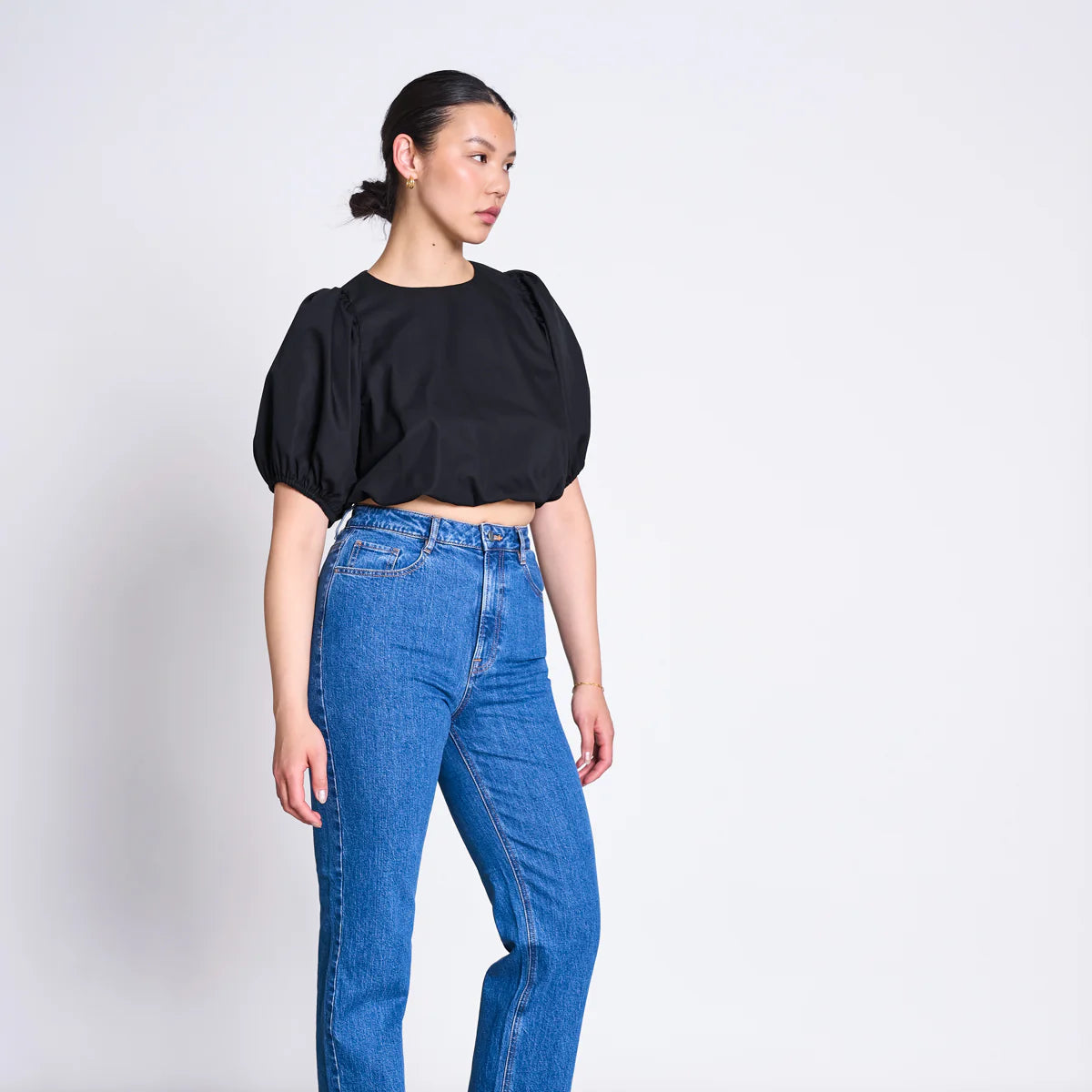 Frau in blauem Jeans und schwarzem, bauchfreiem Oberteil steht seitlich vor einem neutralen Hintergrund.