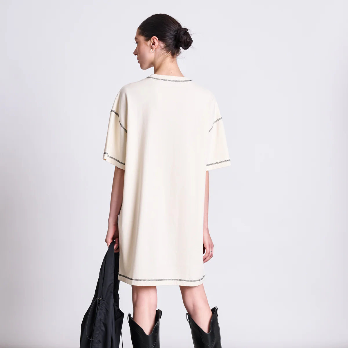 Eine Person mit dunklen Haaren in einem Dutt trägt ein lockeres, kurzärmeliges, beigefarbenes Kleid und hohe schwarze Stiefel. Sie steht mit dem Rücken zur Kamera und hält eine schwarze Tasche an ihrer Seite vor einem schlichten Hintergrund.