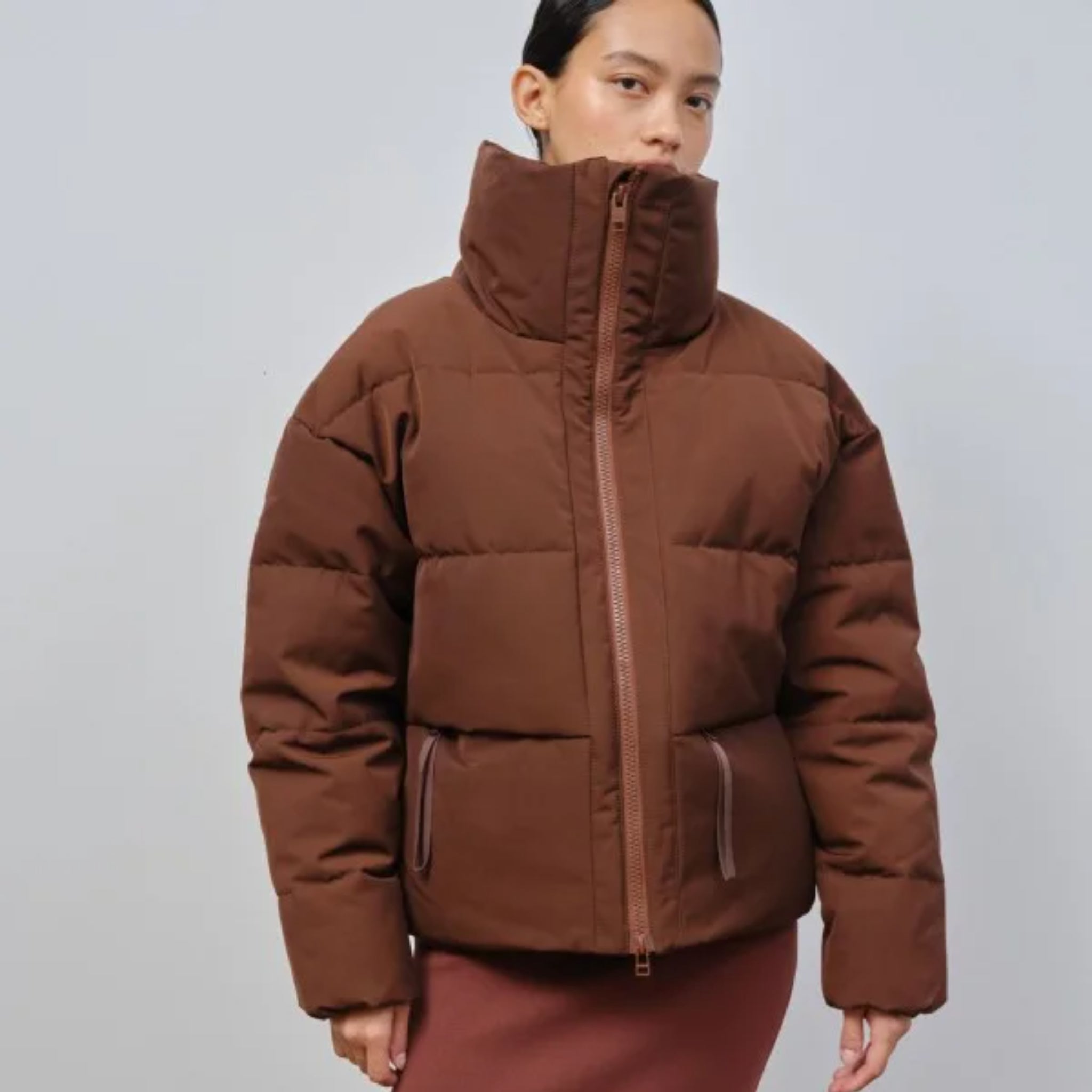 Person mit brauner Steppjacke und hohem Kragen vor neutralem Hintergrund.