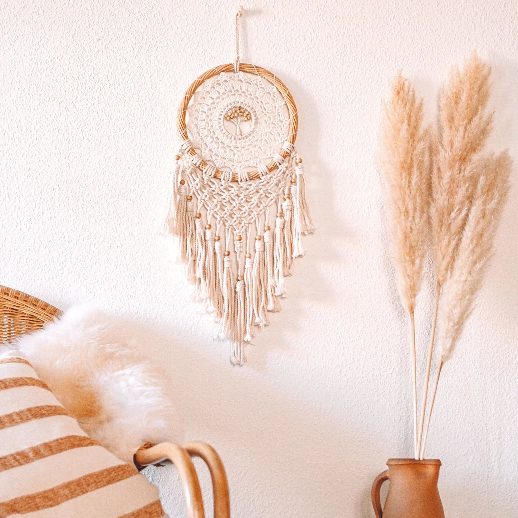 Ein beiger Makramee-Wandbehang hängt über einem Stuhl mit einem gestreiften Kissen und einer Kunstfelldecke. Daneben steht hohes Pampasgras in einer braunen Vase vor einer hellen Wand und schafft eine gemütliche Boho-Deko-Szene.