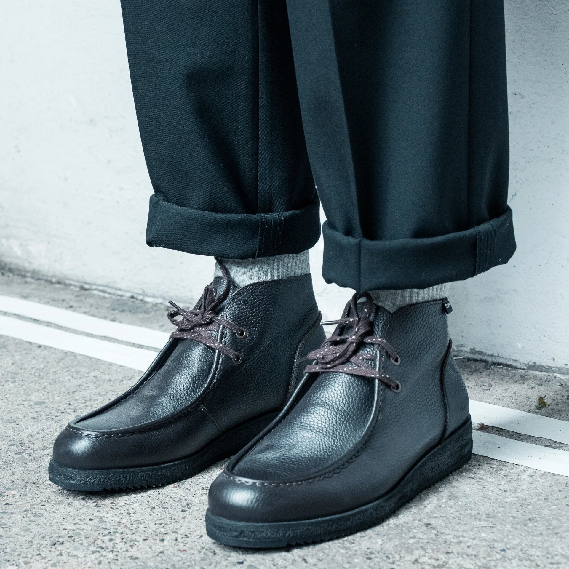 Eine Person, die schwarze Lederschnürstiefel, weiße Socken und eine schwarze Hose mit Bündchen trägt, steht auf einer Betonfläche in der Nähe einer weißen Wand.