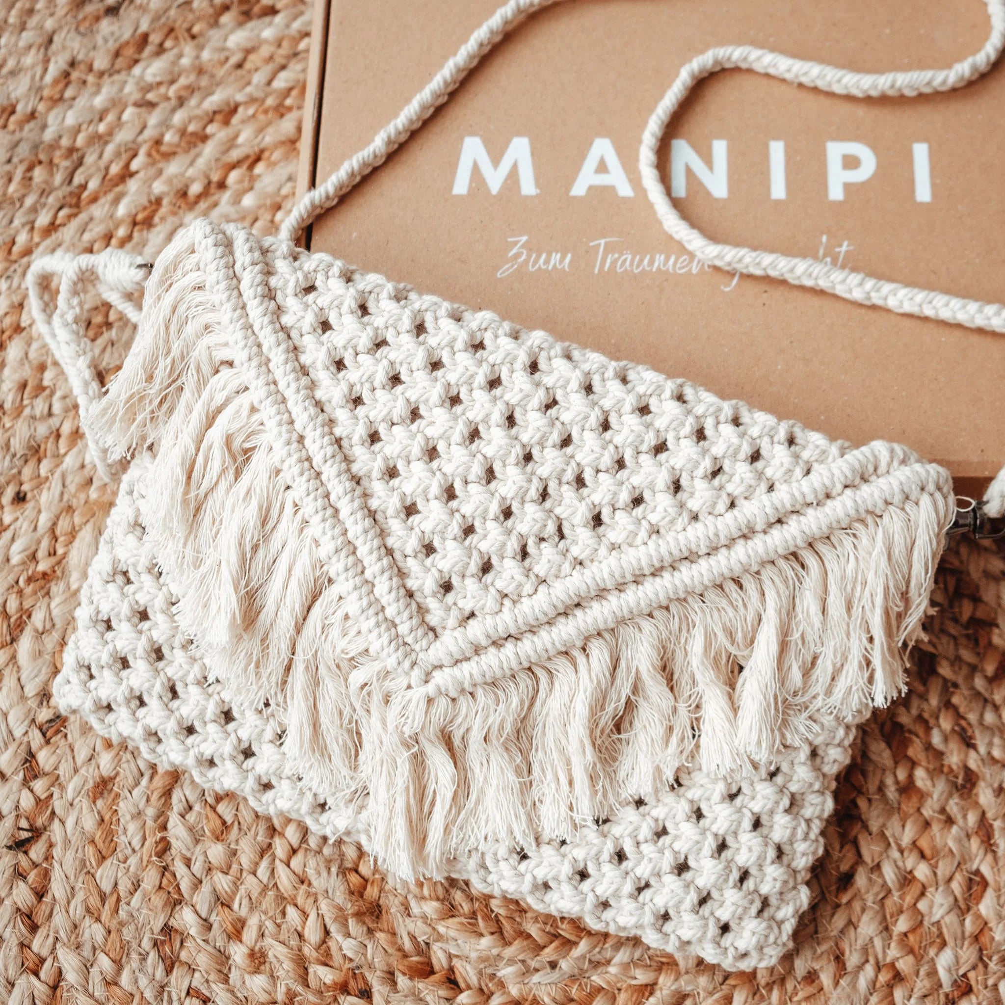 Eine cremefarbene, gewebte Makramee-Clutch-Tasche mit Fransen an der Klappe liegt auf einer strukturierten Webmatte. Dahinter befindet sich eine braune Schachtel mit der Aufschrift MANIPI. Die Tasche hat einen langen Riemen aus demselben Material.