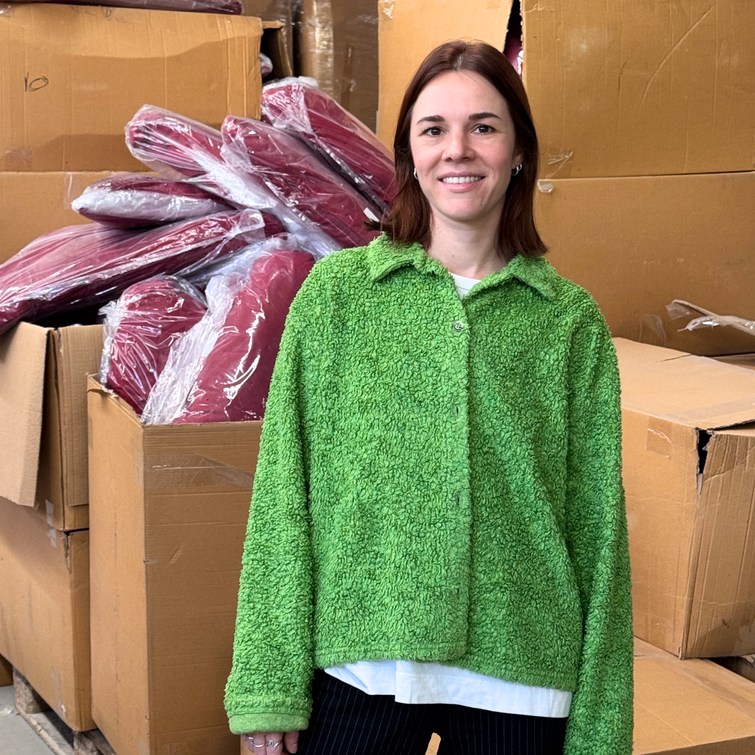 Eine lächelnde Person in einer grünen Jacke steht vor großen Kartons und gestapelten gepolsterten Bezügen.
