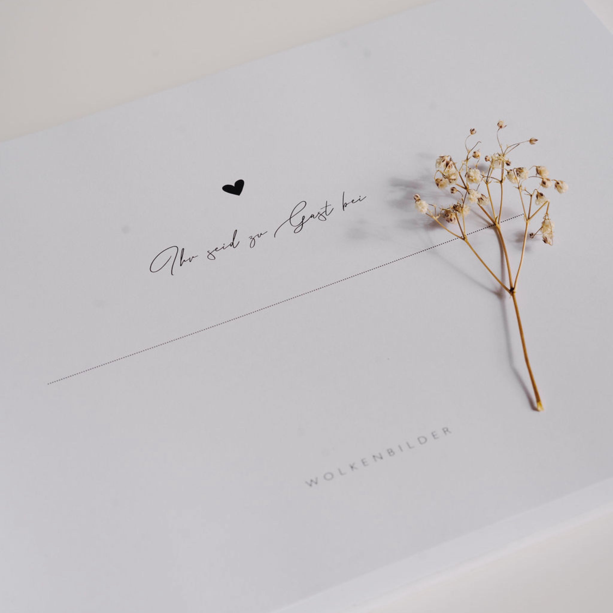 Eine weiße Karte mit der Aufschrift "Ihr seid zu Gast bei" neben einem kleinen, getrockneten Blumenstrauß.