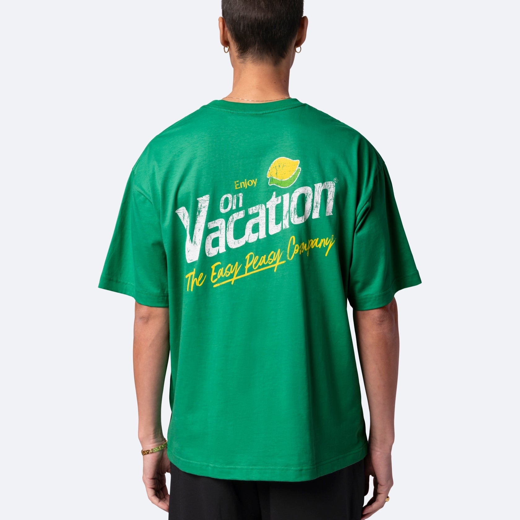 Eine Person, die ein grünes T-Shirt mit gelbem und weißem Text auf der Rückseite trägt, auf dem "Enjoy on Vacation" steht, mit einer Zitronengrafik und "The Easy Peasy Company" darunter, steht vor einem schlichten hellen Hintergrund.