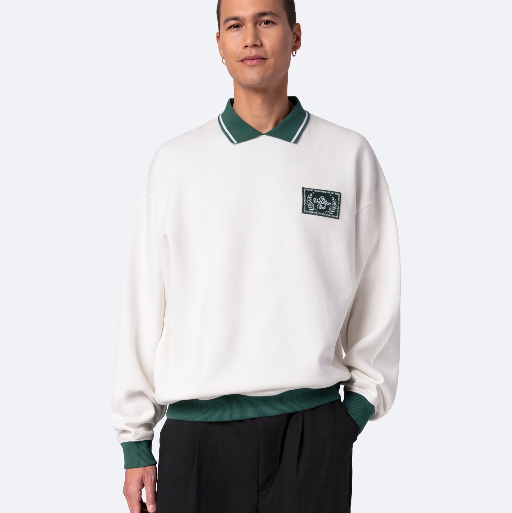Eine Person, die ein weißes Sweatshirt mit grünem Kragen und grünen Manschetten und einem grünen gestickten Aufnäher auf der Brust trägt, steht mit einer Hand in der Tasche vor einem schlichten hellen Hintergrund.
