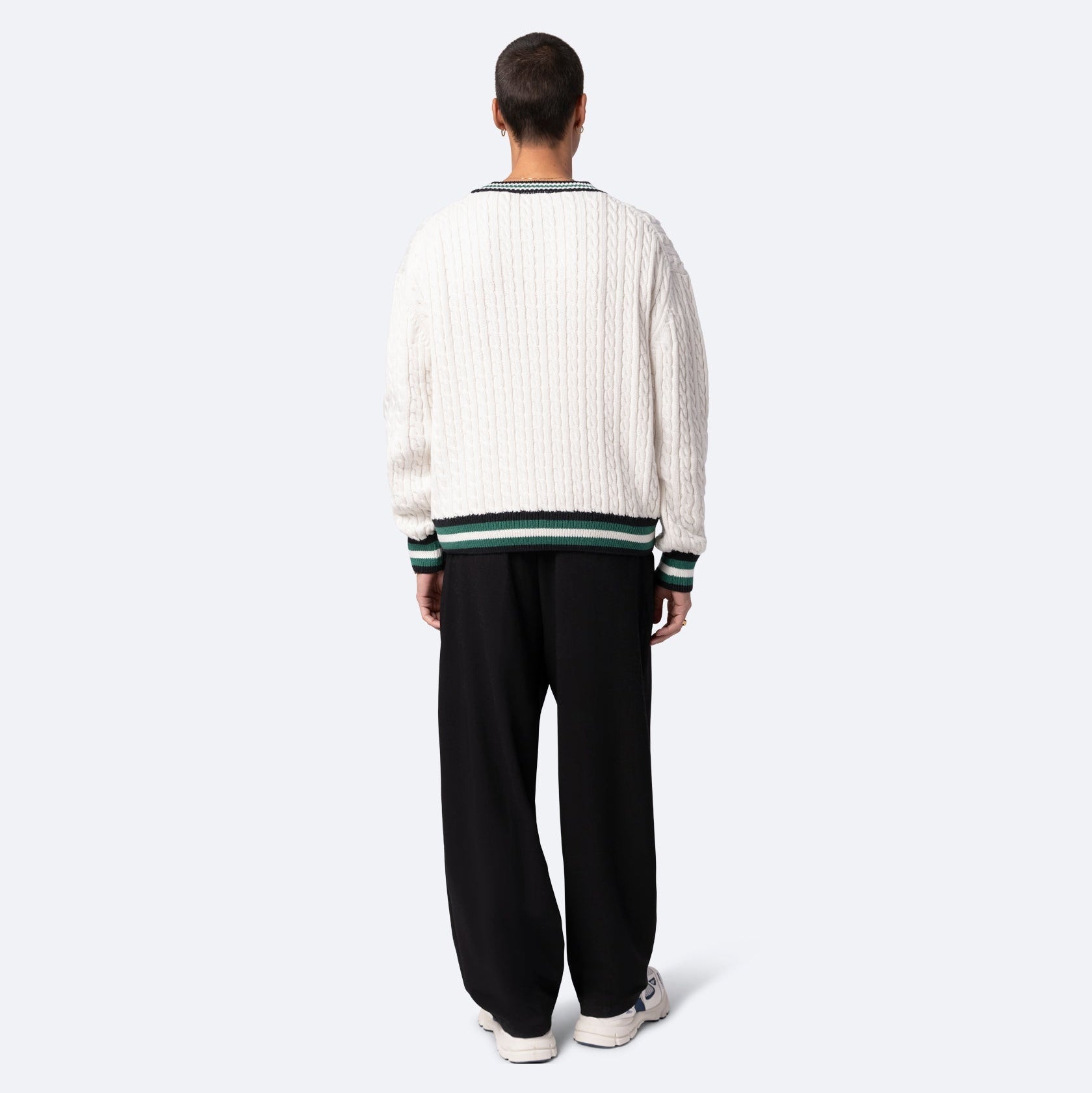 Eine Person steht mit dem Gesicht zur Wand und trägt einen weißen Zopfstrickpullover mit grünem und schwarzem Rand, eine weite schwarze Hose und weiße Turnschuhe vor einem schlichten hellgrauen Hintergrund.
