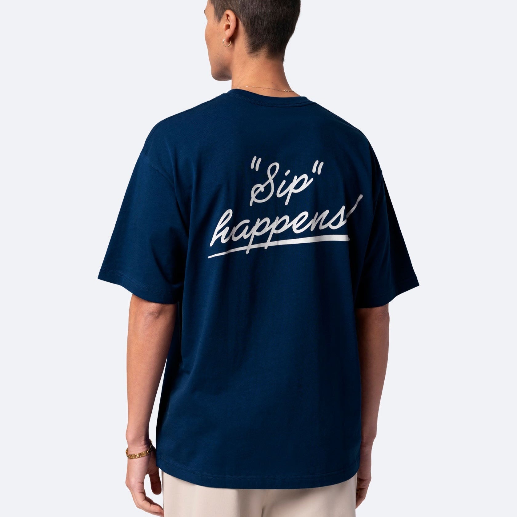 Eine Person steht mit dem Gesicht zur Wand und trägt ein marineblaues T-Shirt mit der Aufschrift "Sip happens!" in weißer Schrift auf dem Rücken. Die Person hat kurze Haare und trägt eine helle Hose.
