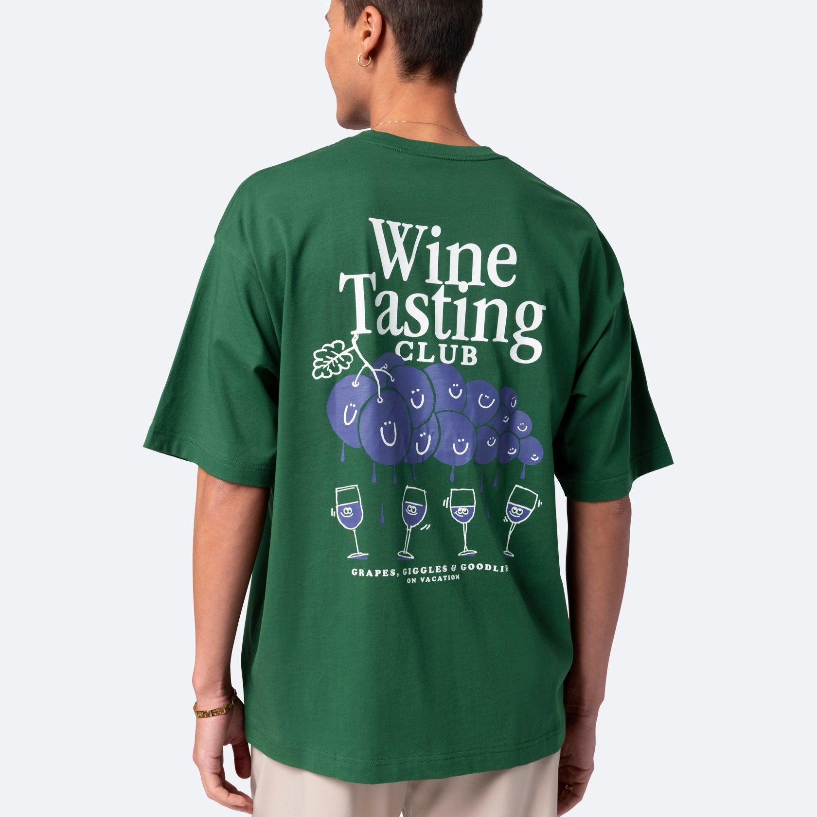 Eine Person, die ein grünes Wine Tasting Club-T-Shirt mit einer Cartoon-Grafik von lächelnden Trauben und Weingläsern auf der Rückseite trägt, steht vor einem schlichten hellen Hintergrund.