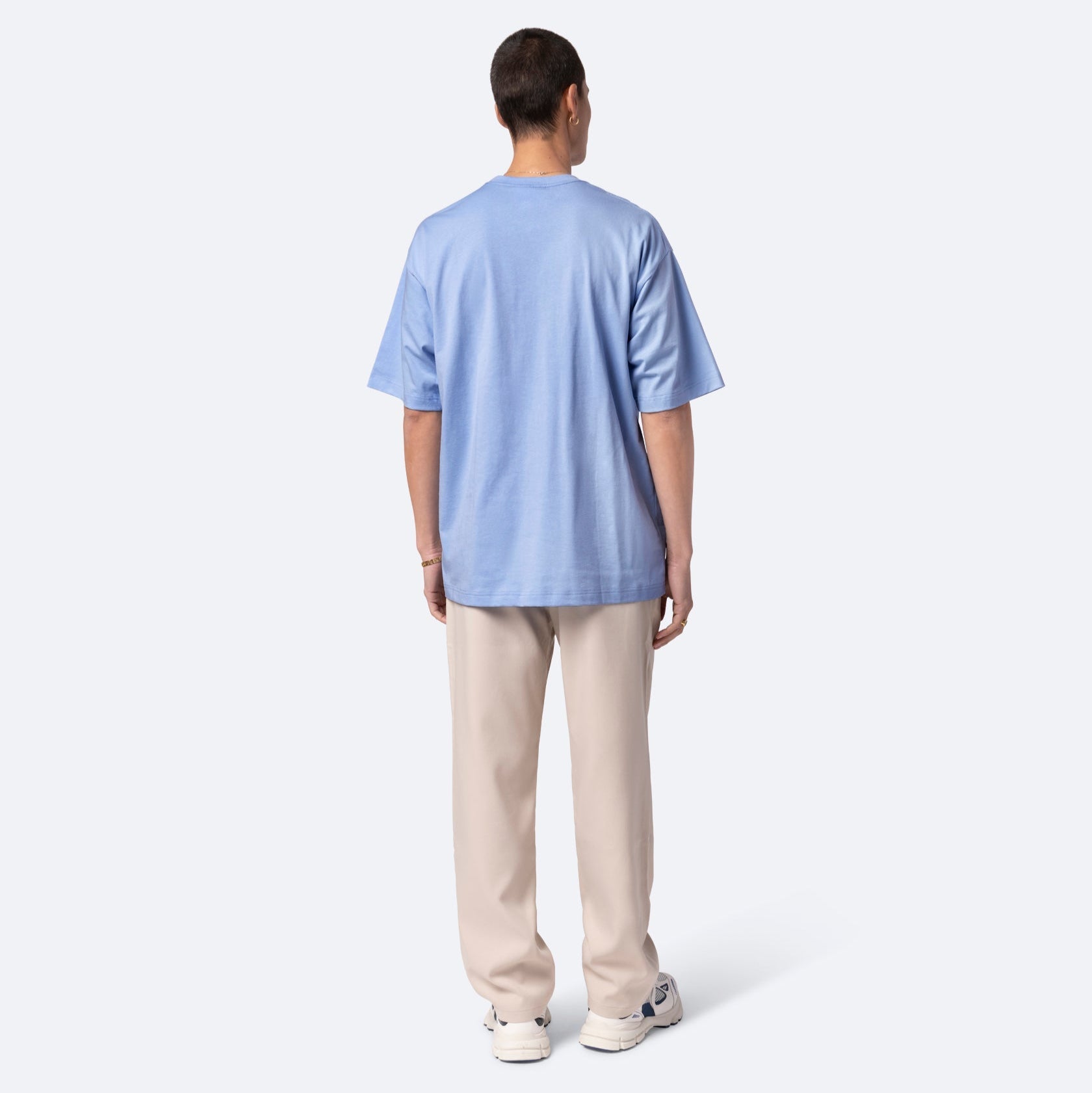 Eine Person steht mit dem Gesicht zur Wand, trägt ein hellblaues T-Shirt, eine beigefarbene Hose und weiße Turnschuhe vor einem schlichten hellen Hintergrund.
