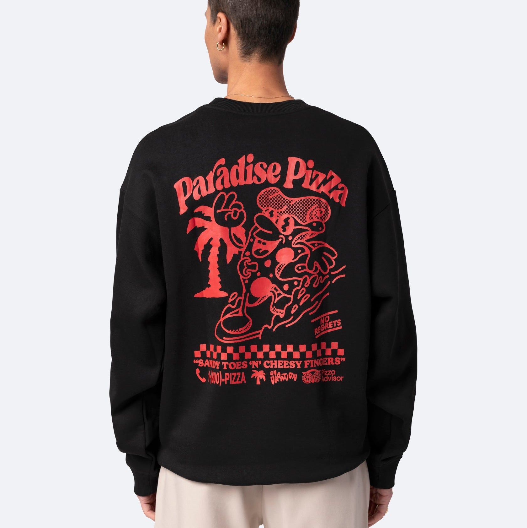 Eine Person mit kurzen Haaren blickt nach vorne und trägt ein schwarzes Sweatshirt mit einer großen roten Grafik auf dem Rücken, auf der "Paradise Pizza" steht und die eine Zeichentrickfigur, eine Palme und ein verspieltes Pizzamotiv zeigt.