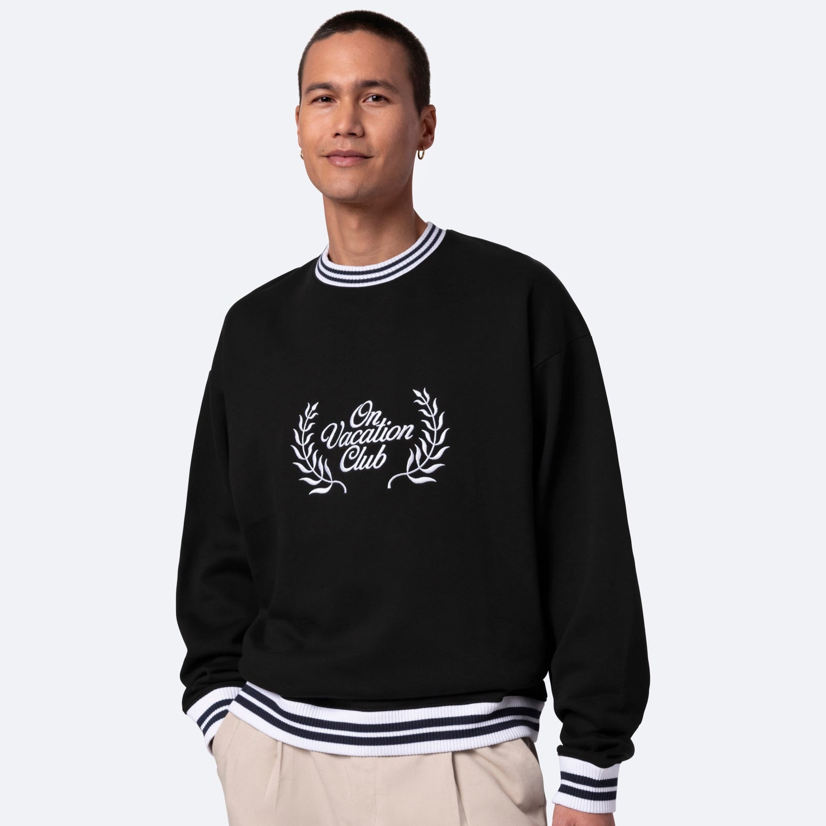 Eine Person, die ein schwarzes Sweatshirt mit weiß gestreiften Manschetten und Kragen, eine beigefarbene Hose und den Text Vacation Club mit aufgedruckten Lorbeerzweigen auf der Vorderseite trägt, steht vor einem schlichten hellen Hintergrund.