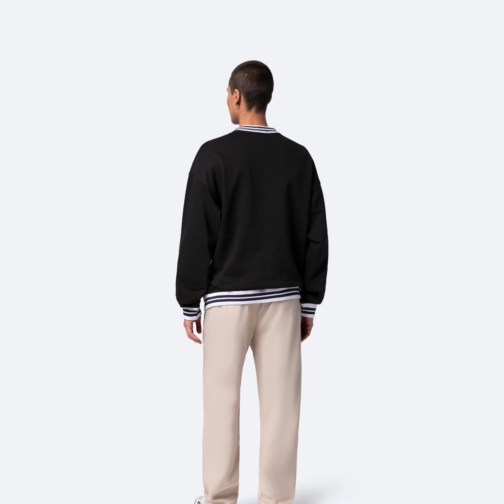 Ein Mann steht mit dem Gesicht zur Wand und trägt einen schwarzen Pullover mit weißen Streifen, eine beigefarbene Hose und weiße Turnschuhe vor einem schlichten hellgrauen Hintergrund.