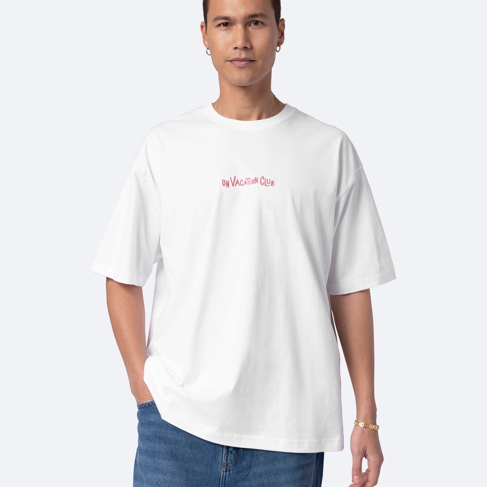 Eine Person, die ein lockeres weißes T-Shirt mit der Aufschrift ON VACATION CLUB in kleinen roten Buchstaben auf der Brust, blaue Jeans, einen Ohrring und ein goldenes Armband trägt, steht vor einem schlichten hellen Hintergrund.