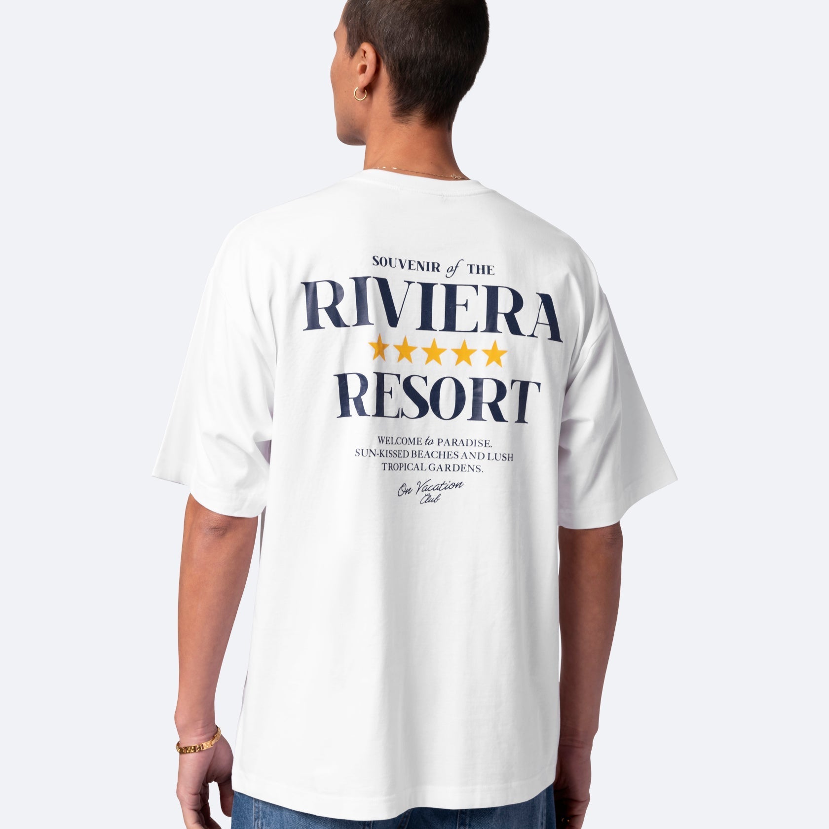 Eine Person trägt ein weißes T-Shirt mit der Aufschrift Souvenir of the Riviera Resort und goldenen Sternen auf dem Rücken, zusammen mit einem kleineren Text und einem Detail im Stil einer Unterschrift darunter. Die Person blickt von der Kamera weg.