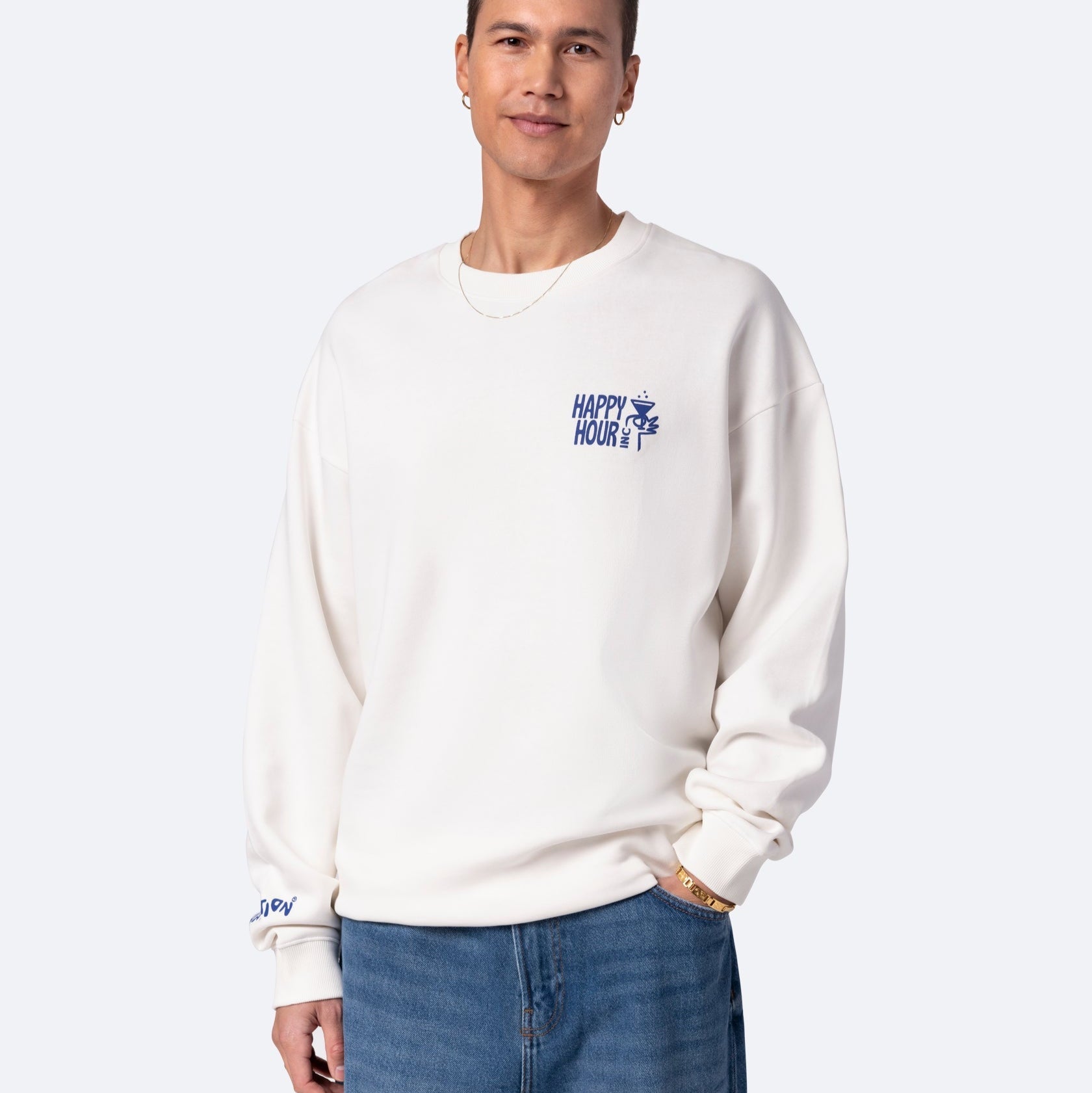Eine Person, die ein weißes Sweatshirt mit der Aufschrift HAPPY HOUR und einer kleinen Grafik auf der Brust, blaue Jeans, eine Goldkette und eine Armbanduhr trägt, steht vor einem hellgrauen Hintergrund, lächelt leicht und hat die Hände in den Taschen.