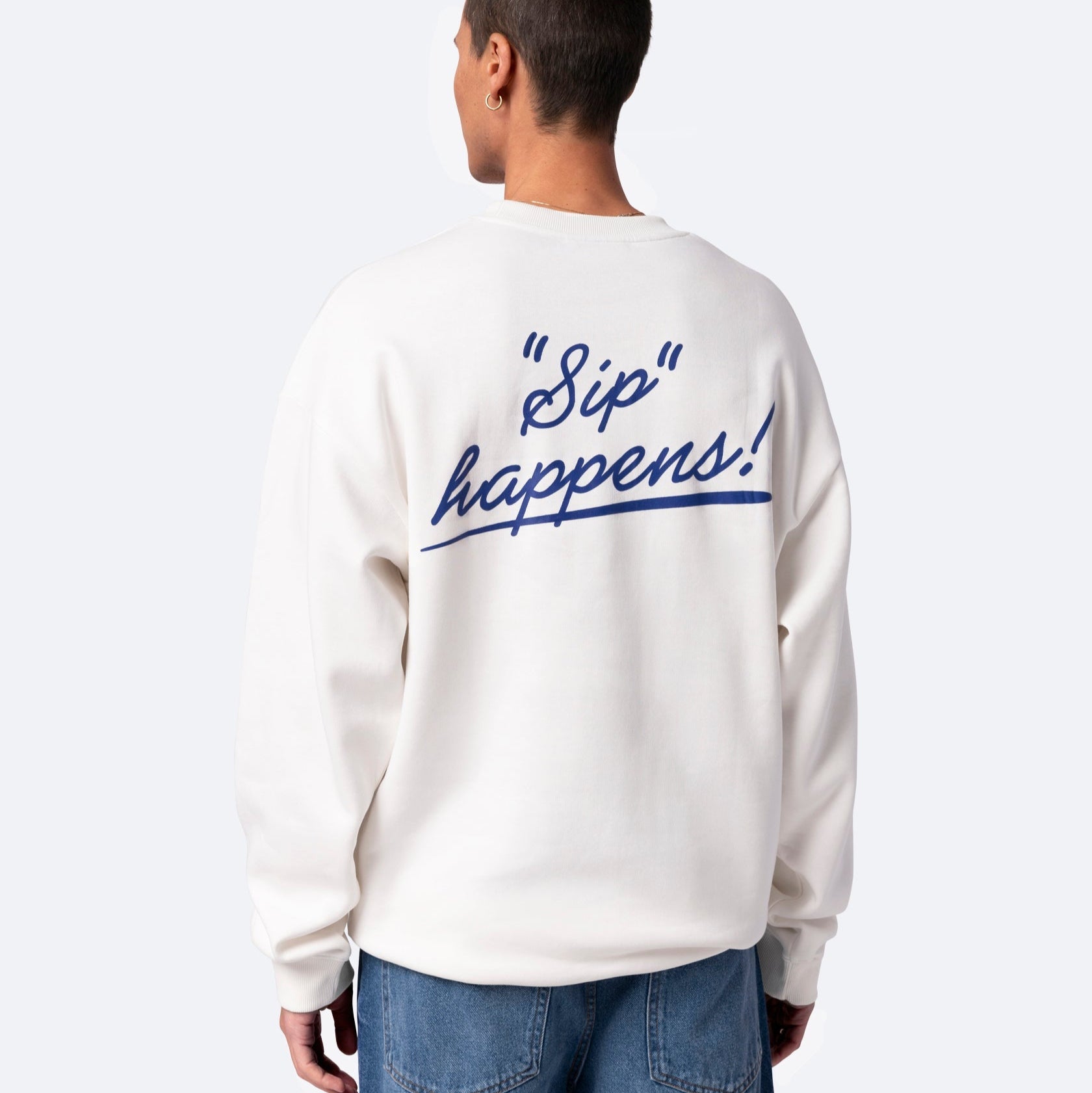 Eine Person, die ein weißes Sweatshirt mit der Aufschrift Sip happens! in blauer Schreibschrift auf dem Rücken trägt, steht mit dem Rücken zur Kamera und trägt eine blaue Jeans.