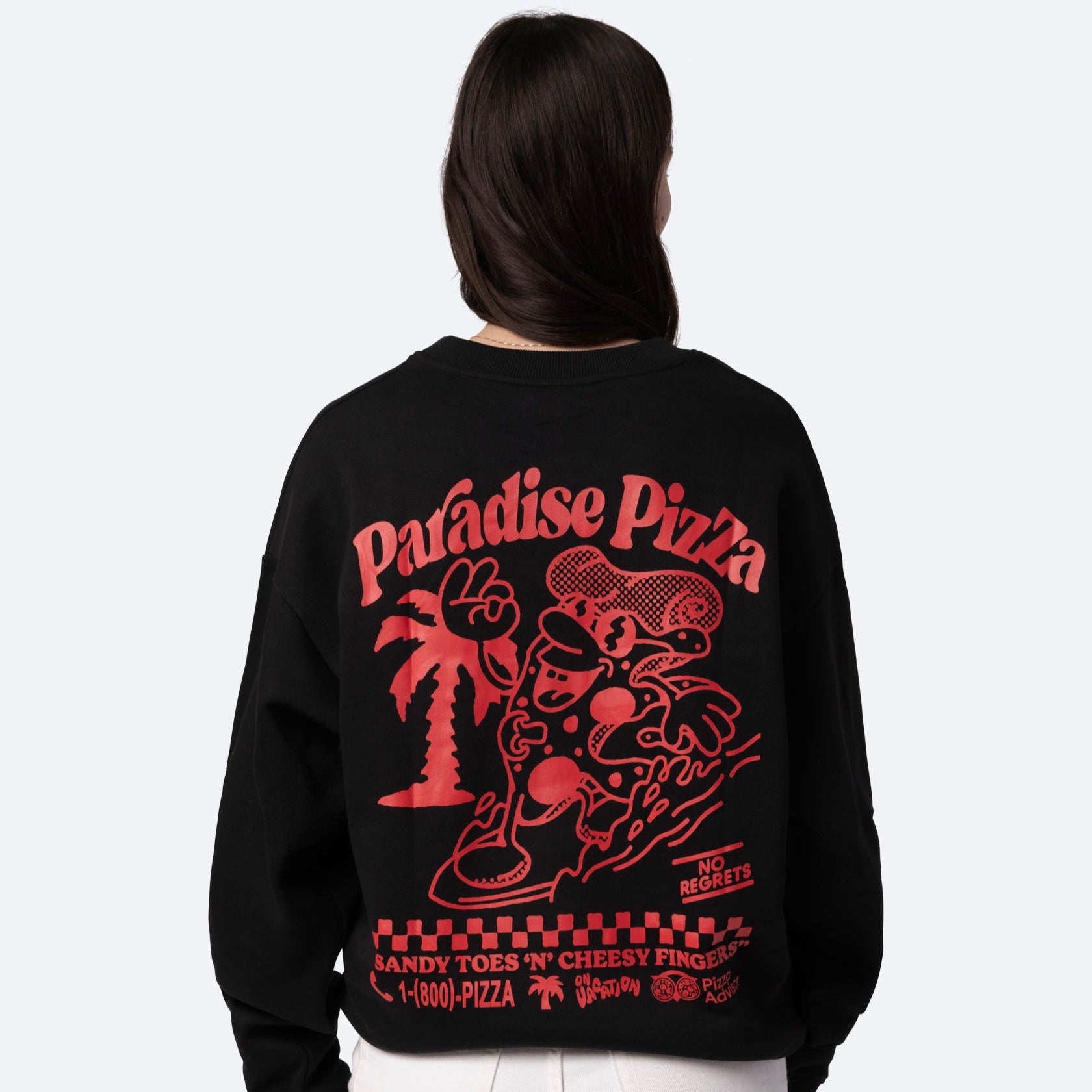 Eine Person, die ein schwarzes Sweatshirt mit einer großen, roten Grafik auf dem Rücken trägt, auf der ein Cartoon-Pizzastück beim Surfen zu sehen ist, eine Palme und der Text "Paradise Pizza" und "Sandy Toes N Cheesy Fingers".
