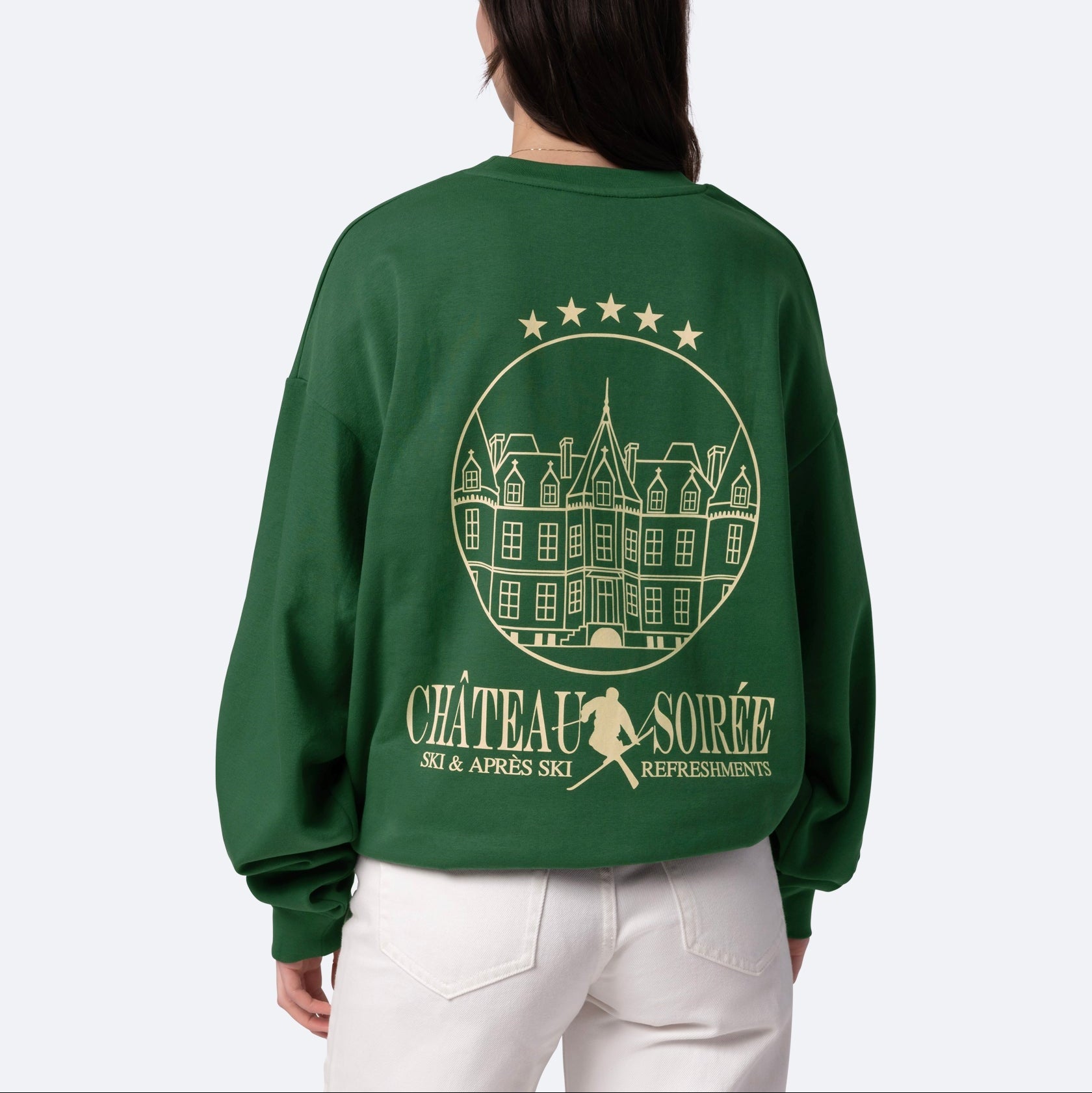 Eine Person trägt ein dunkelgrünes Sweatshirt mit einer goldenen Strichzeichnung eines Schlosses, fünf Sternen und dem Text CHÂTEAU SOIRÉE Ski & Après Ski Refreshments auf dem Rücken. Die Person schaut von sich weg.