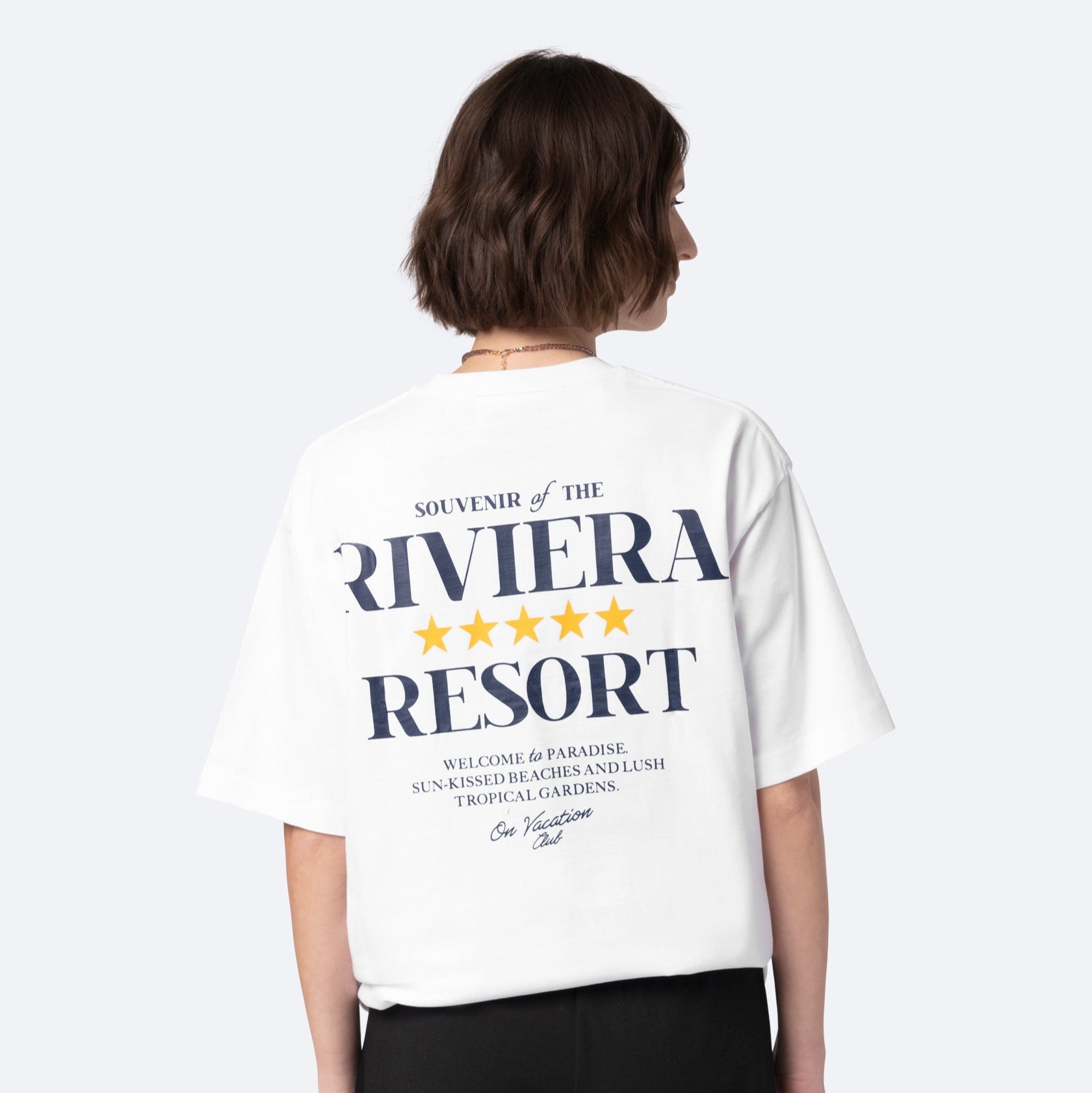 Eine Person mit kurzen braunen Haaren trägt ein weißes T-Shirt mit der großen Aufschrift "Souvenir of the Riviera Resort" auf der Rückseite, gefolgt von einem kleineren dekorativen Text und gelben Sternen. Die Person blickt von der Kamera weg.