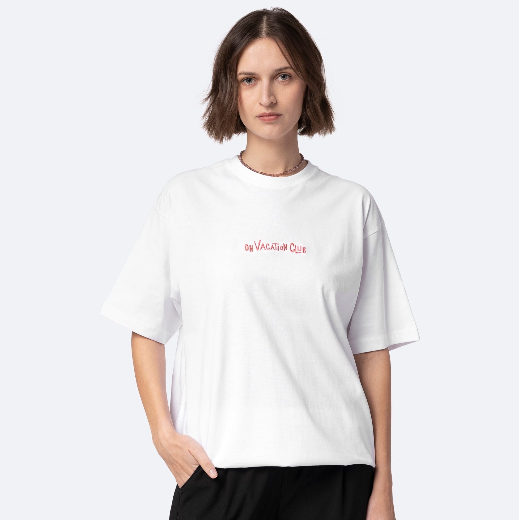 Eine Frau mit kurzen braunen Haaren trägt ein lockeres weißes T-Shirt mit der Aufschrift ON VACATION CLUB in kleinen roten Buchstaben und eine schwarze Hose. Sie steht mit einer Hand in der Tasche vor einem schlichten hellen Hintergrund.