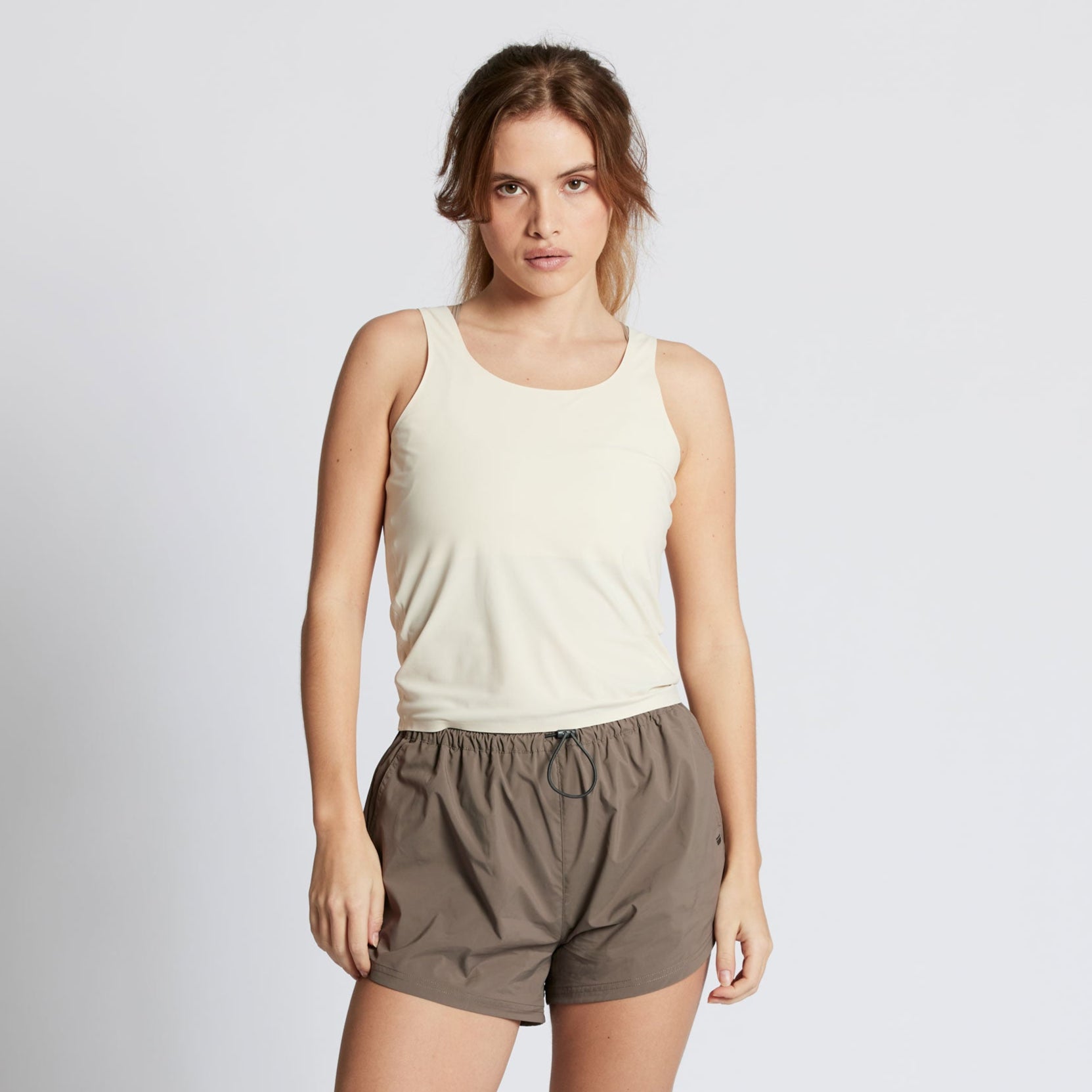 Eine Frau mit langen braunen Haaren, die ein ärmelloses beiges Oberteil und weite graue Shorts trägt, steht vor einem schlichten hellen Hintergrund und schaut mit neutralem Blick direkt in die Kamera.