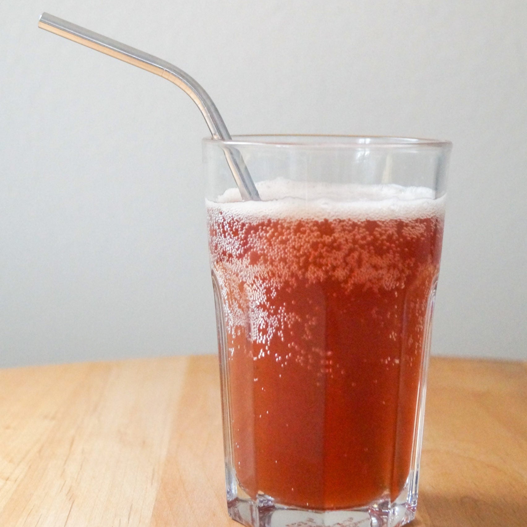 Ein Glas mit sprudelndem rotem Getränk auf einem Holztisch, mit einem Metallstrohhalm darin.