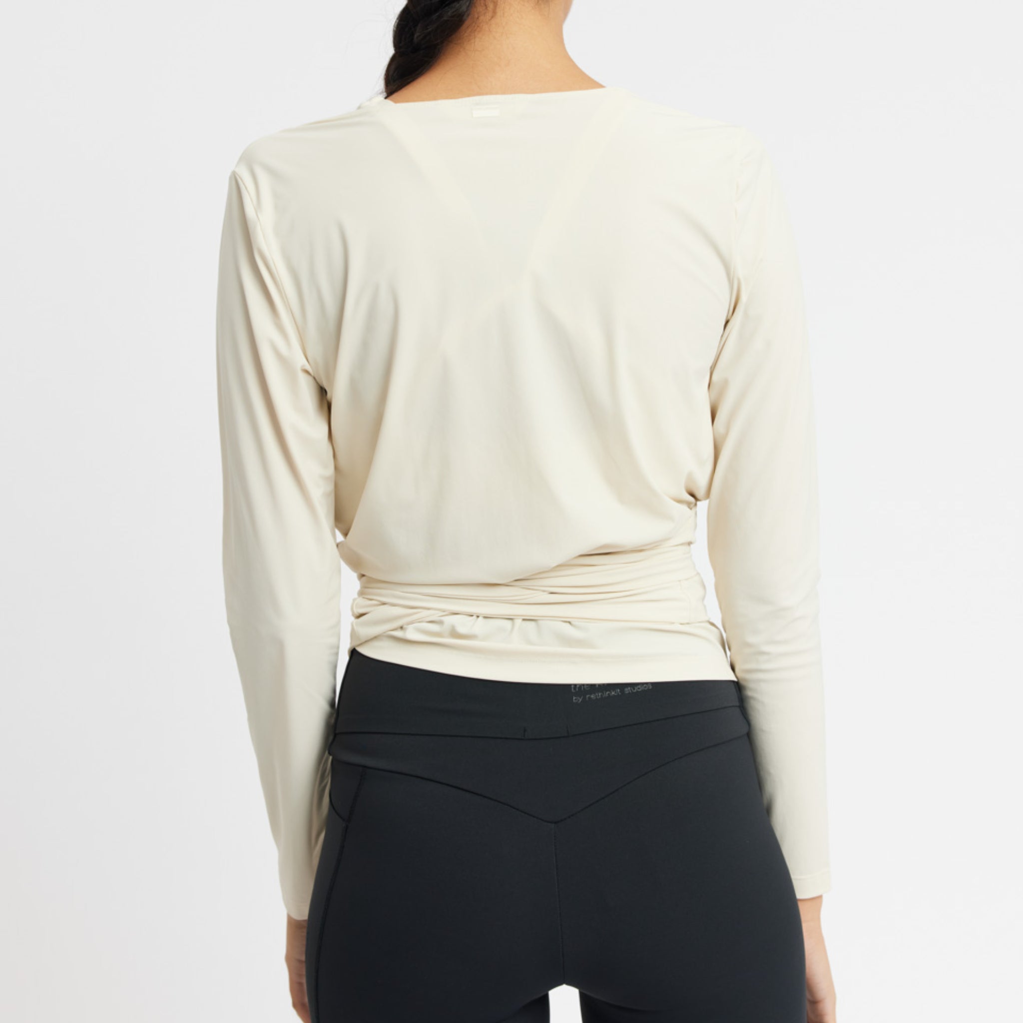 Rückansicht einer Person in einem cremefarbenen Langarmshirt und schwarzen Leggings vor weißem Hintergrund.