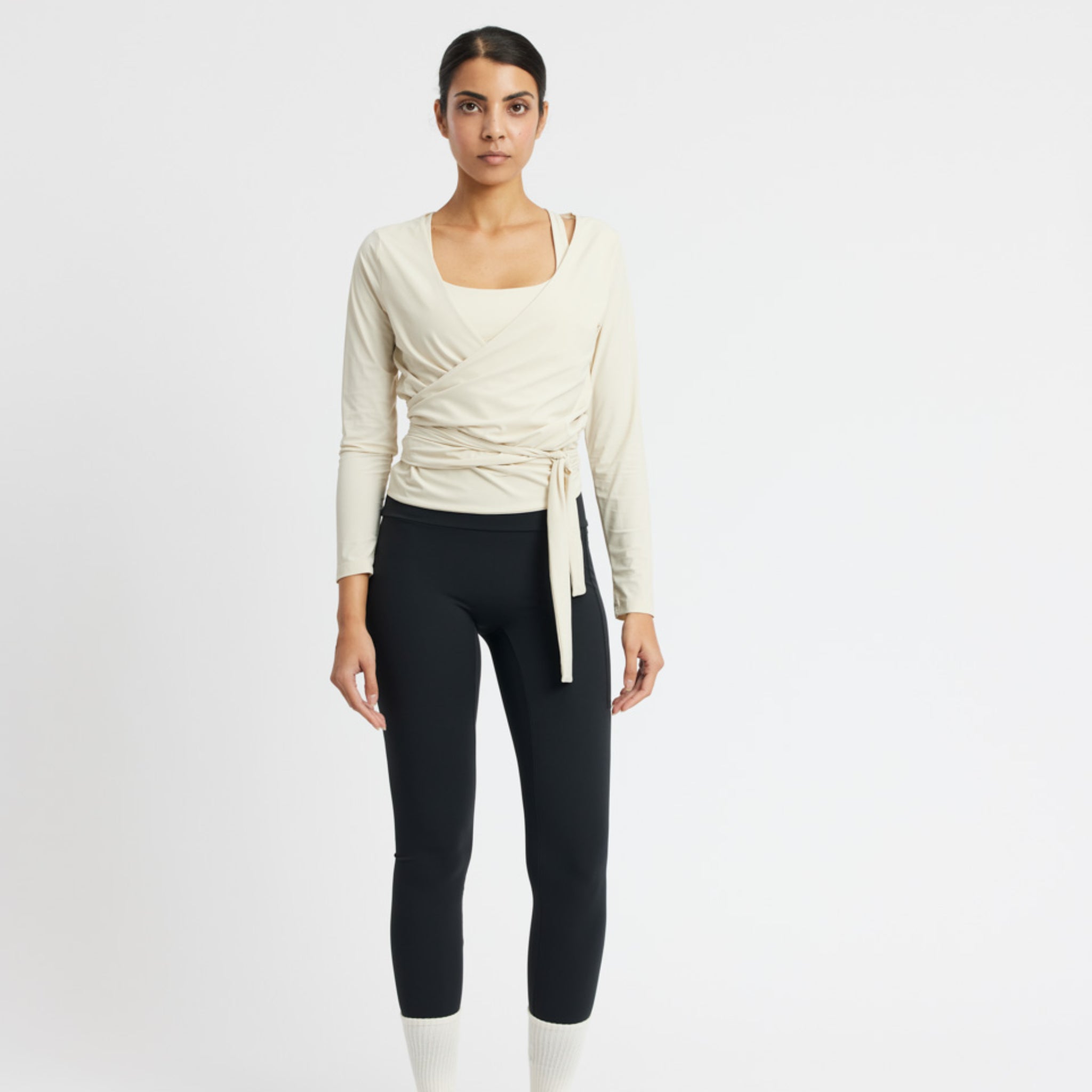 Frau in beiger Wickelbluse und schwarzer Leggings posiert vor einem weißen Hintergrund.