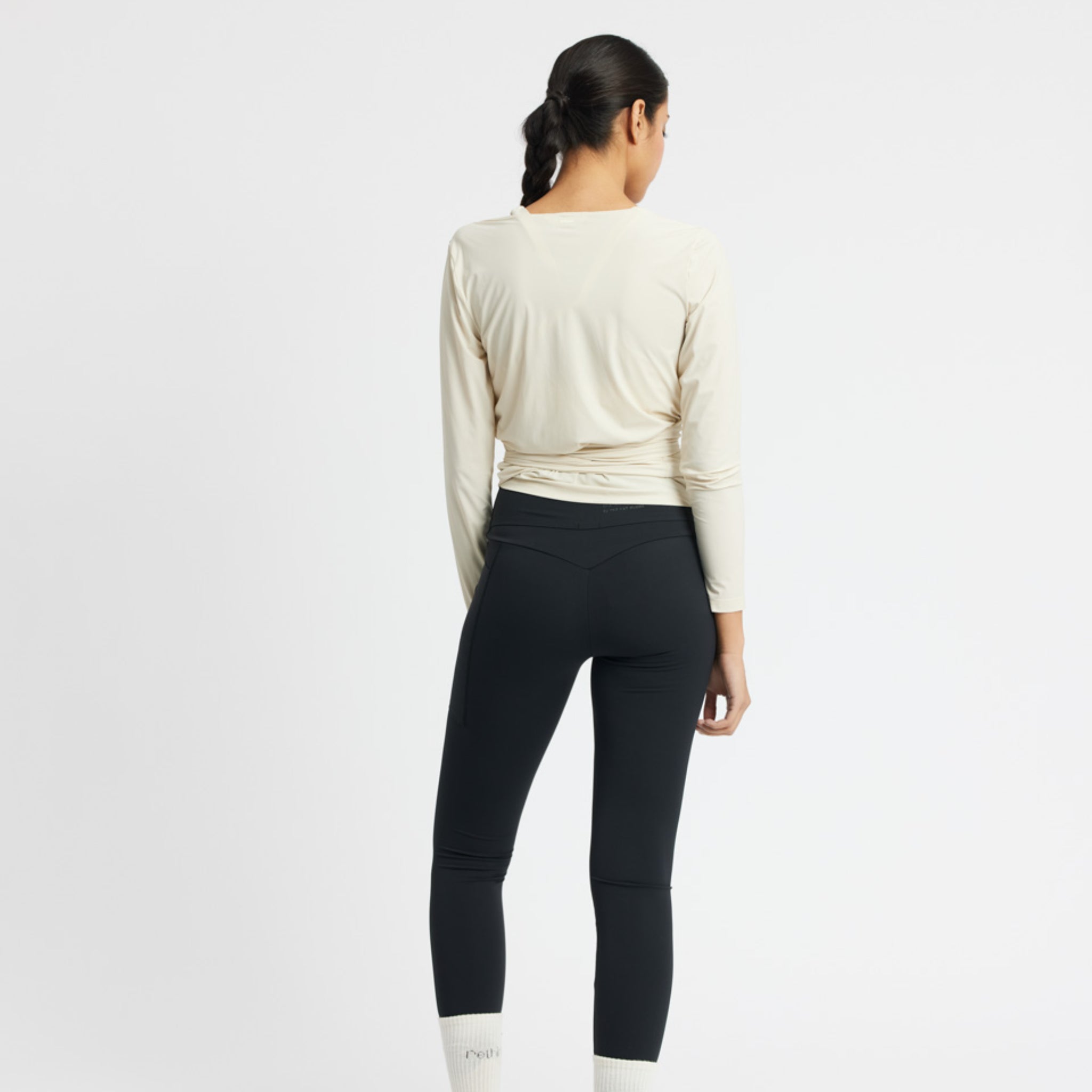 Eine Person in einem beigen Oberteil und schwarzen Leggings steht mit dem Rücken zur Kamera auf einem weißen Hintergrund.