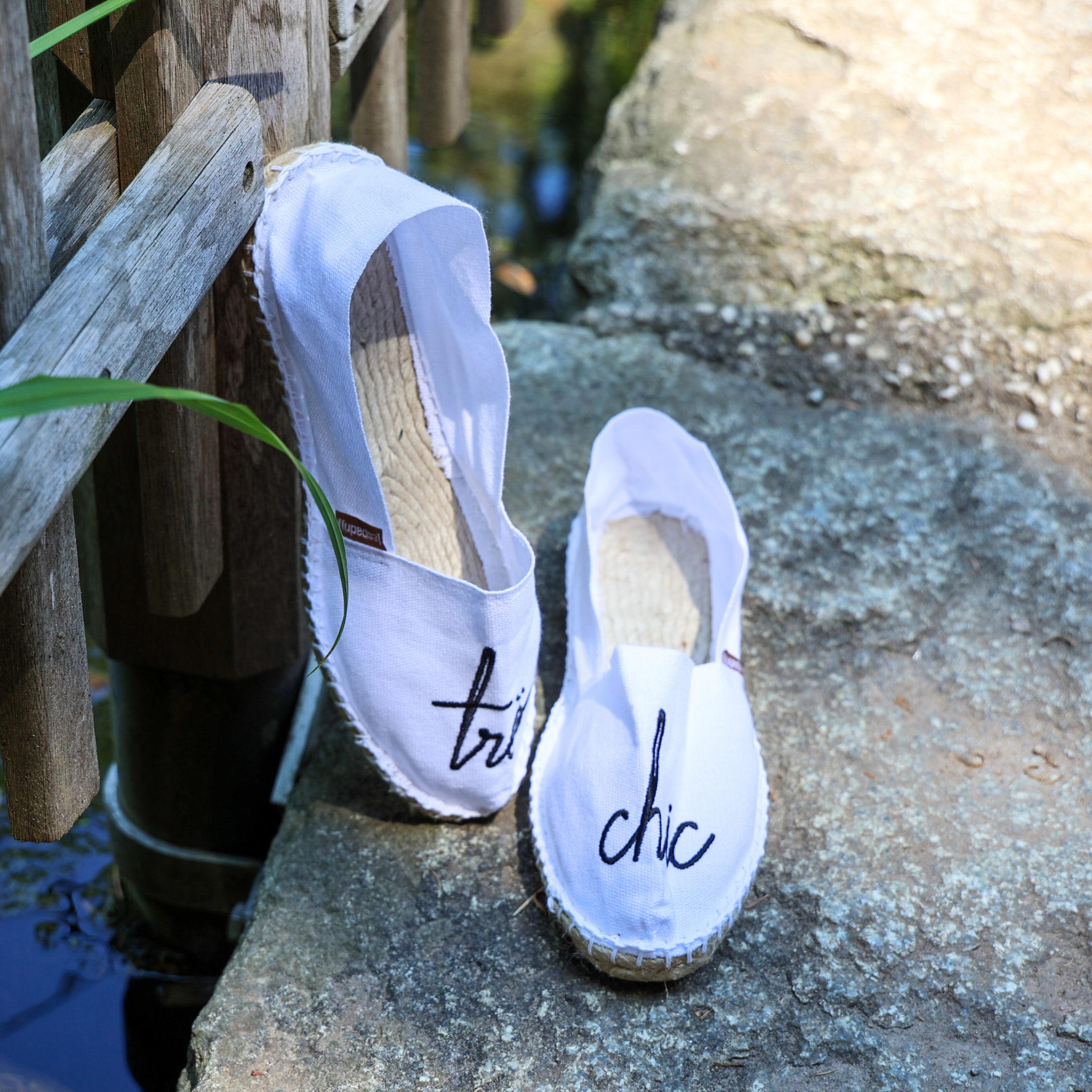 Ein Paar weiße Espadrilles mit schwarzer Stickerei "très" und "chic" auf einer Steinfläche neben einem Holzzaun und einem Teich.
