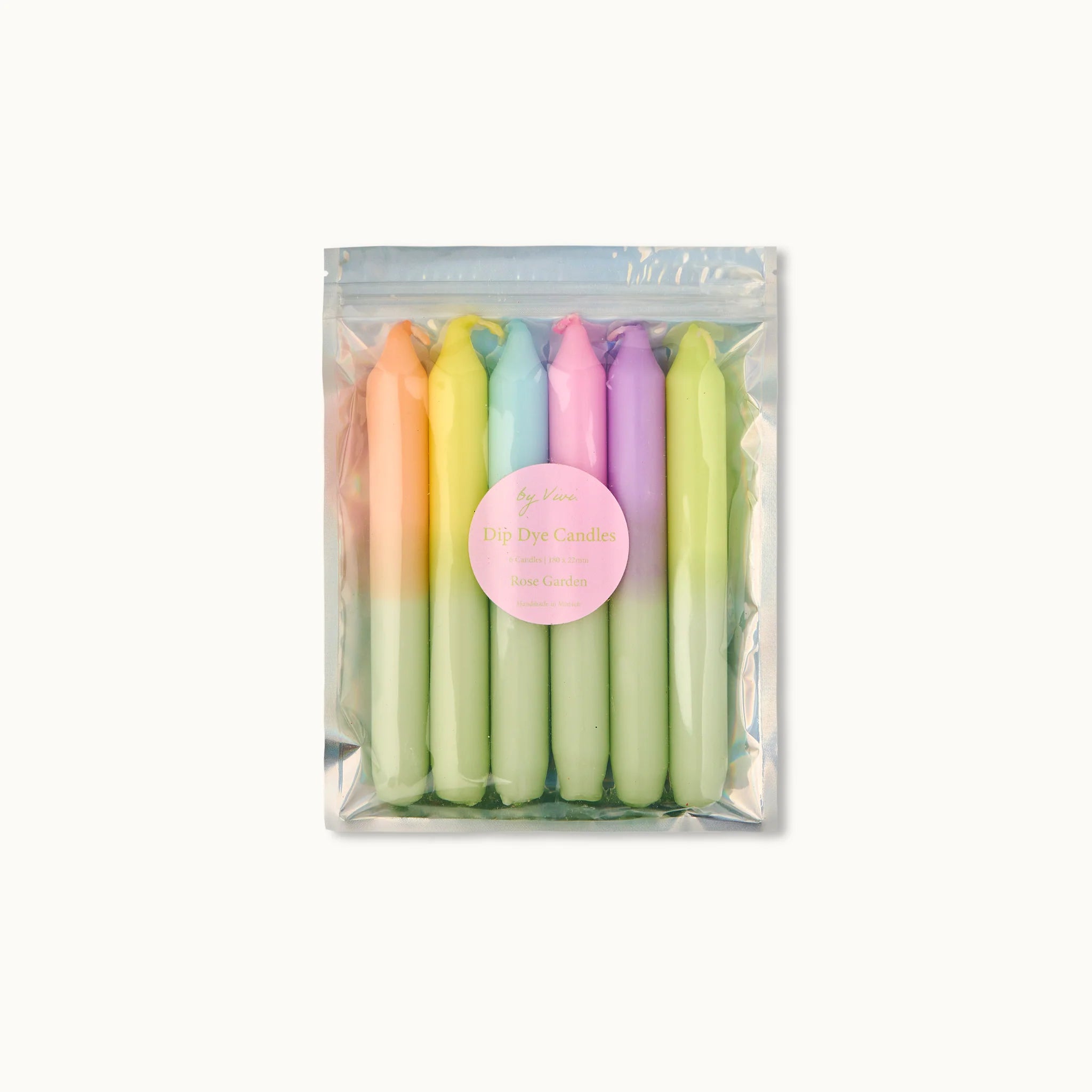 Eine durchsichtige Plastiktüte enthält sechs pastellfarbene Dip-Dye-Kerzen, wobei jede Kerze zwei sanfte Farbtöne mischt. Ein rundes rosafarbenes Etikett auf der Tüte trägt die Aufschrift "Dip Dye Candles" mit zusätzlichem Text darunter. Der Hintergrund ist einfarbig weiß.