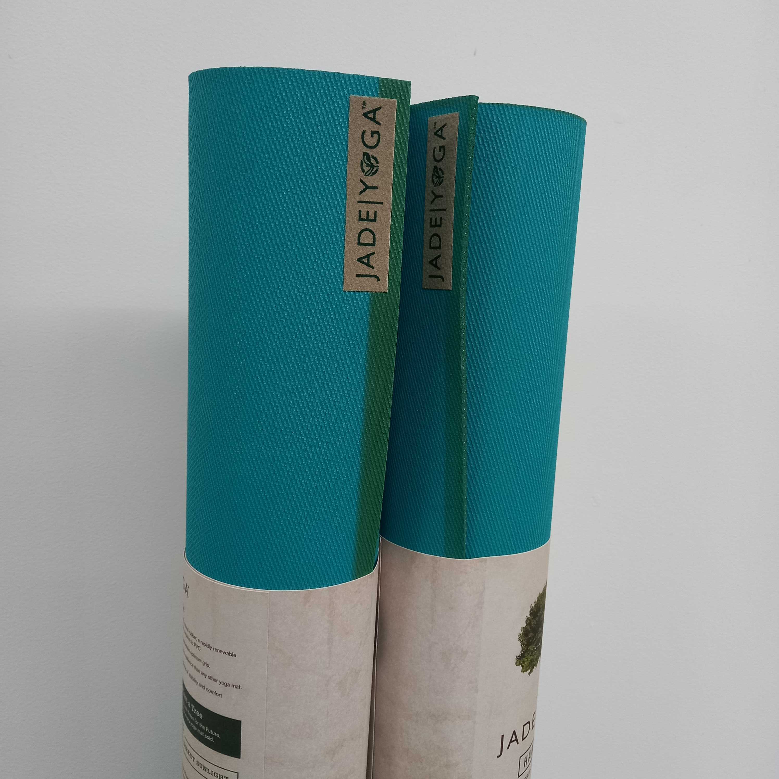 Zwei zusammengerollte, türkisfarbene Yoga-Matten mit dem Etikett "JADE YOGA" stehen nebeneinander an einer Wand.
