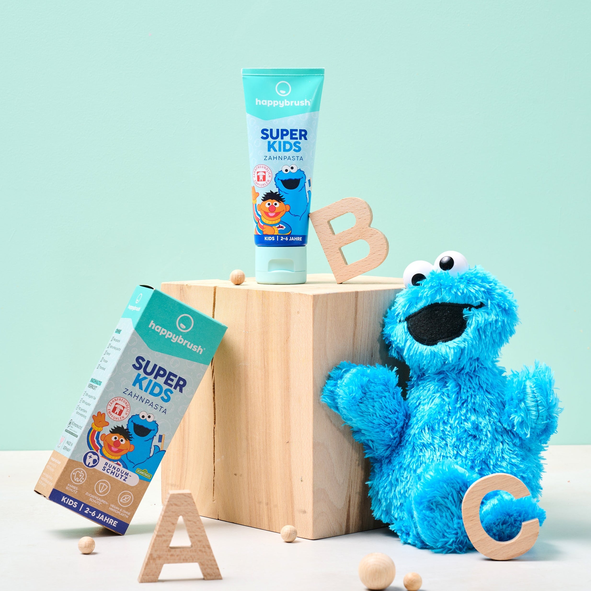 Zahnpasta „Super Kids“ von Happybrush auf einem Holzblock mit blauen Stofftier. Buchstaben A, B, C drumherum.