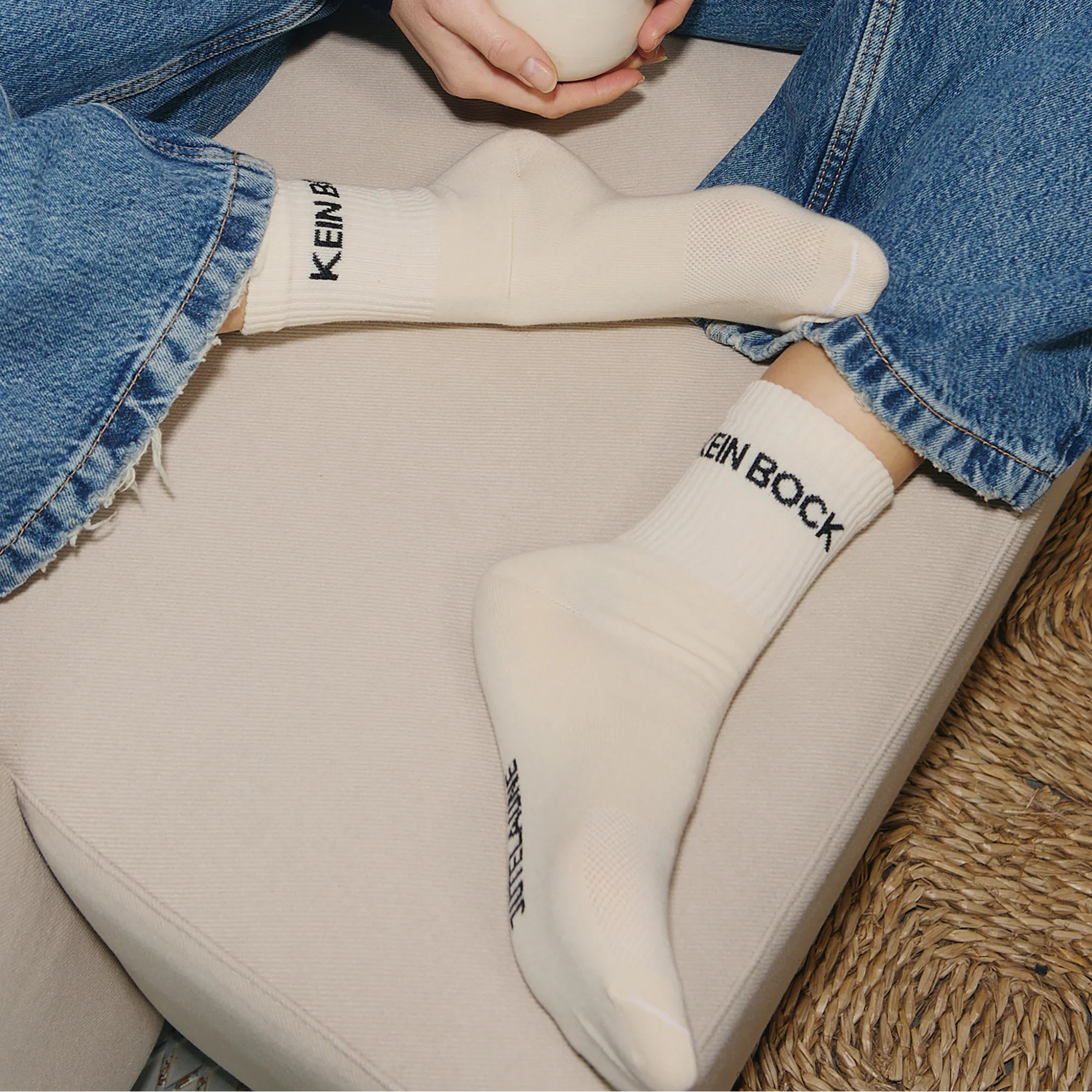 Person in weißen Socken mit der Aufschrift KEIN BOCK in schwarzer Schrift, im Schneidersitz auf einer beigen Couch sitzend, bekleidet mit blauen Jeans, in der Hand eine weiße Tasse. Auf dem Boden ist ein Teil eines gewebten Teppichs zu sehen.