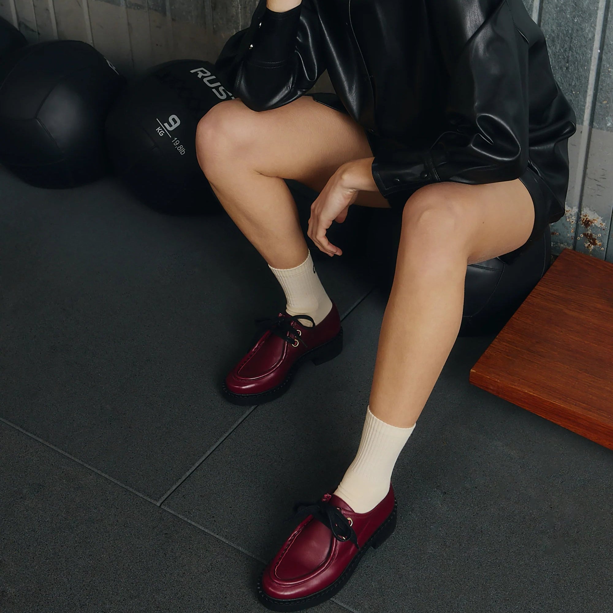 Eine Person in schwarzer Lederjacke, cremefarbenen Socken und weinroten Halbschuhen sitzt auf einem schwarzen Gymnastikball in einem Fitnessstudio, die Beine gekreuzt und mit dem Gesicht außer Sicht. Im Hintergrund sind Fitnessgeräte zu sehen.