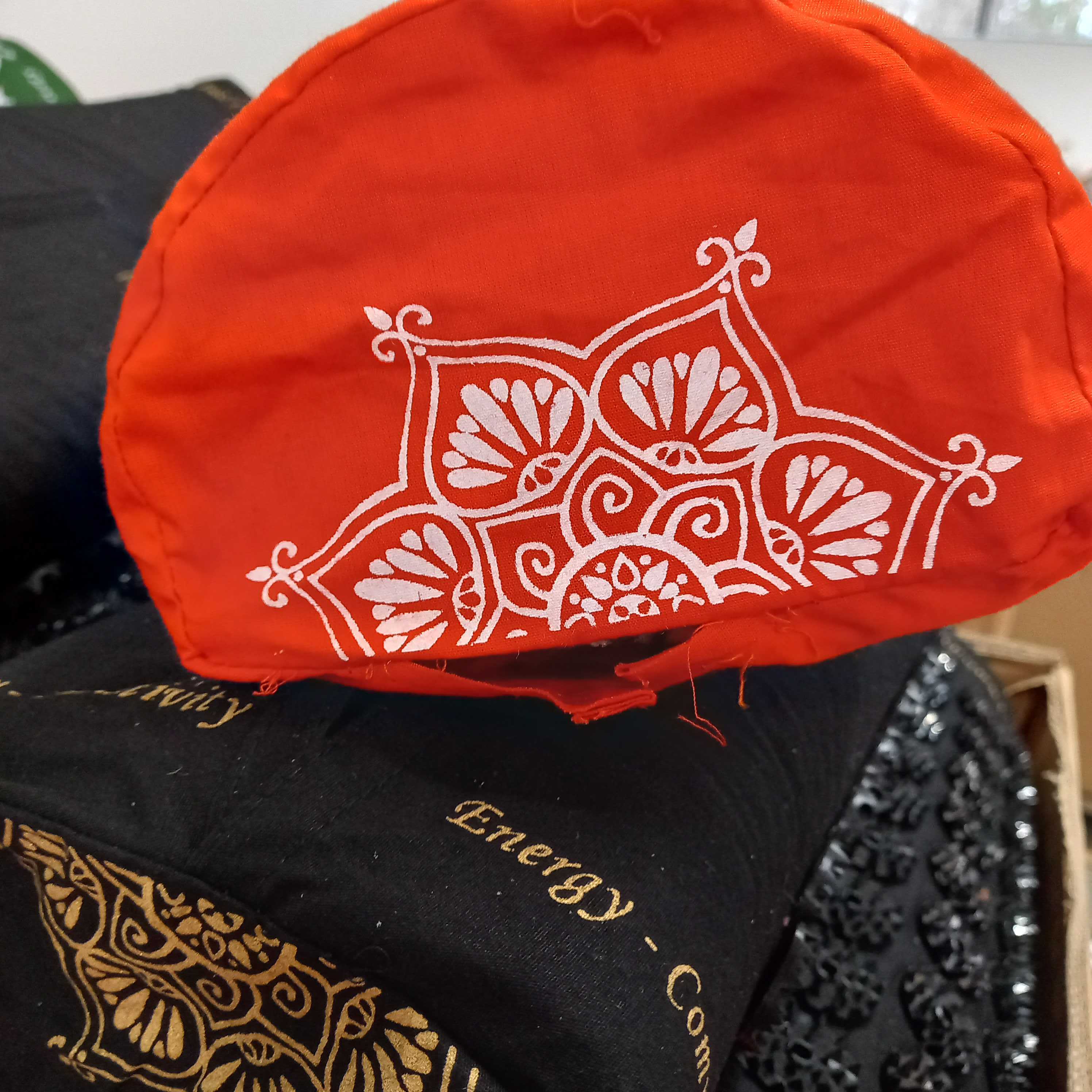 Rote Mütze mit weißem Mandalamuster liegt auf schwarzen Kleidungsstücken mit goldener Stickerei und Text.