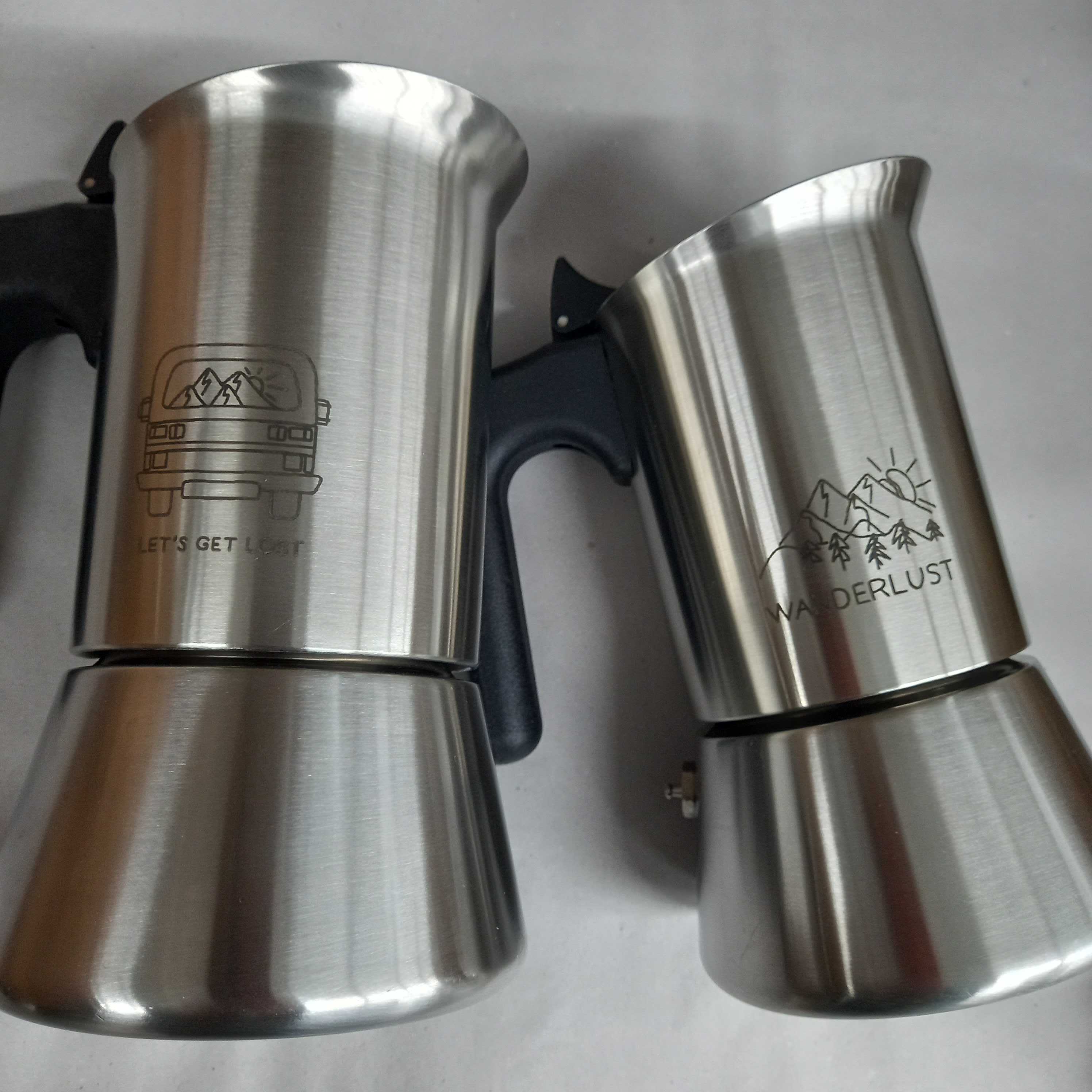 Zwei silberne Espressokocher mit schwarzen Griffen, dekoriert mit Bergmotiven und den Texten "LET'S GET LOST" und "WANDERLUST".