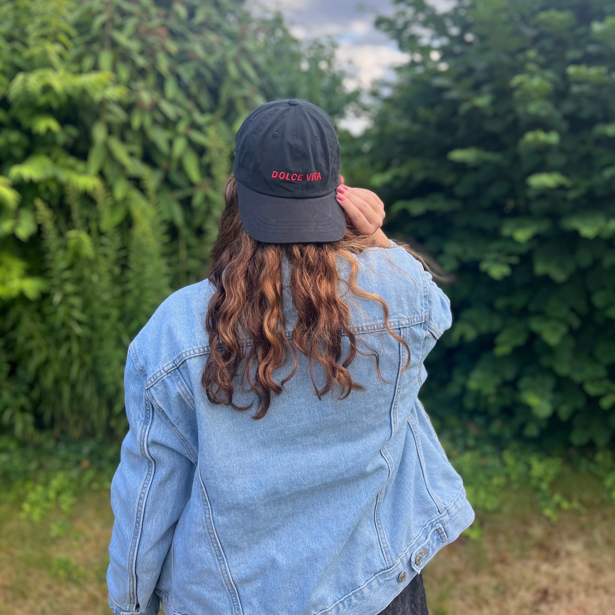 Person mit langer, welliger Haaren trägt eine schwarze Mütze mit Aufschrift "Dolce Vita" und eine Jeansjacke, im Grünen.
