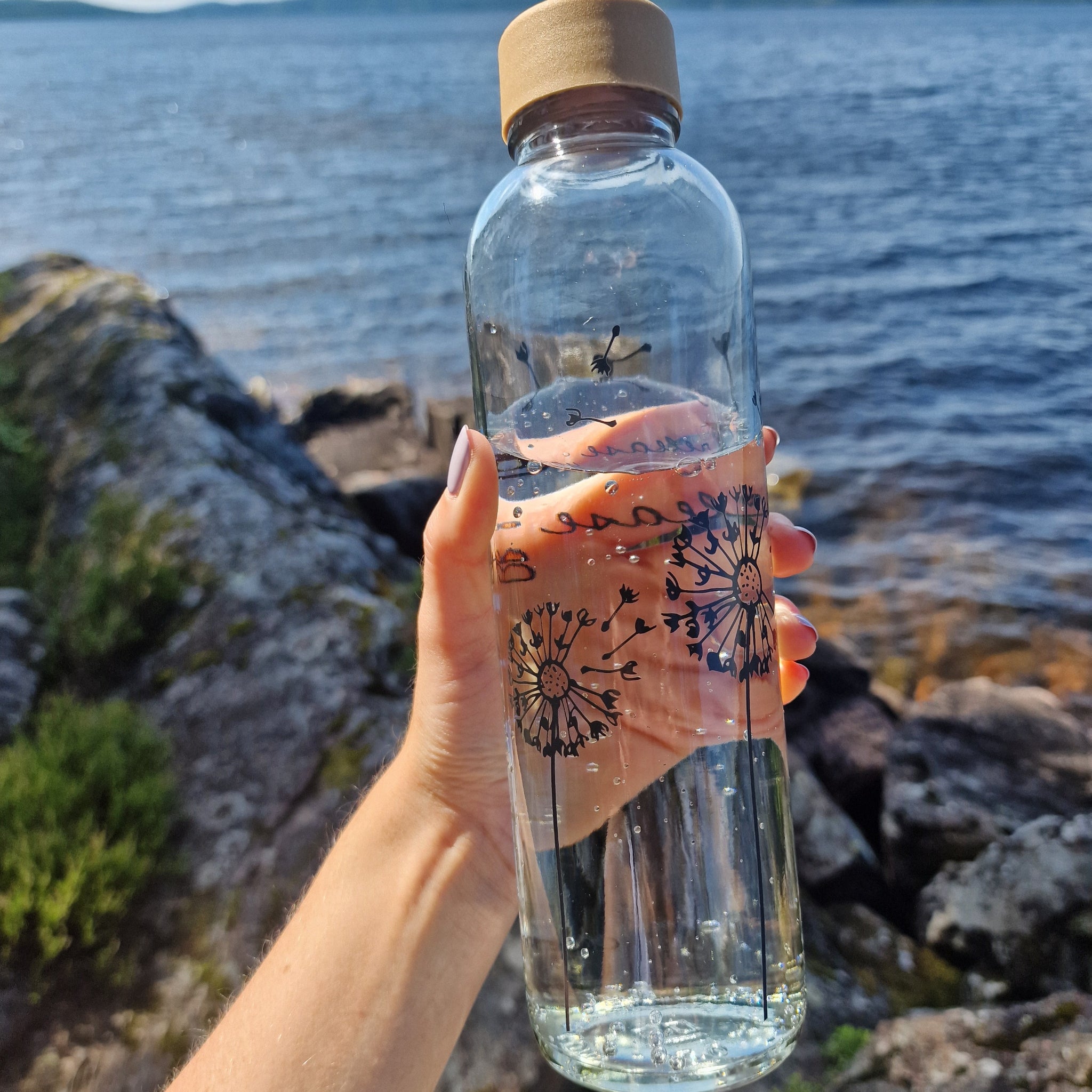 Eine Hand hält eine Glasflasche mit Löwenzahnmuster, im Hintergrund ein See und felsige Küste.