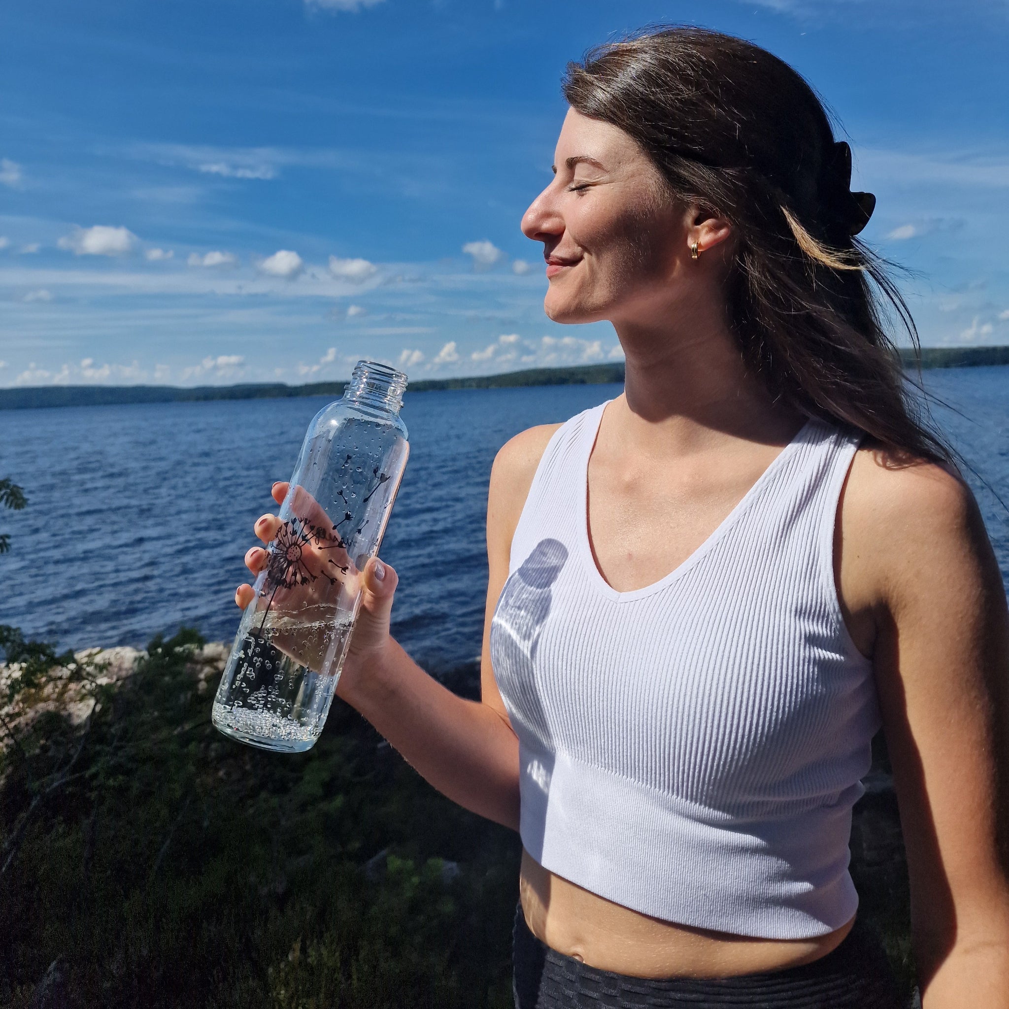 Frau in weißem Top hält lächelnd eine Wasserflasche in der Sonne am See mit blauem Himmel im Hintergrund.