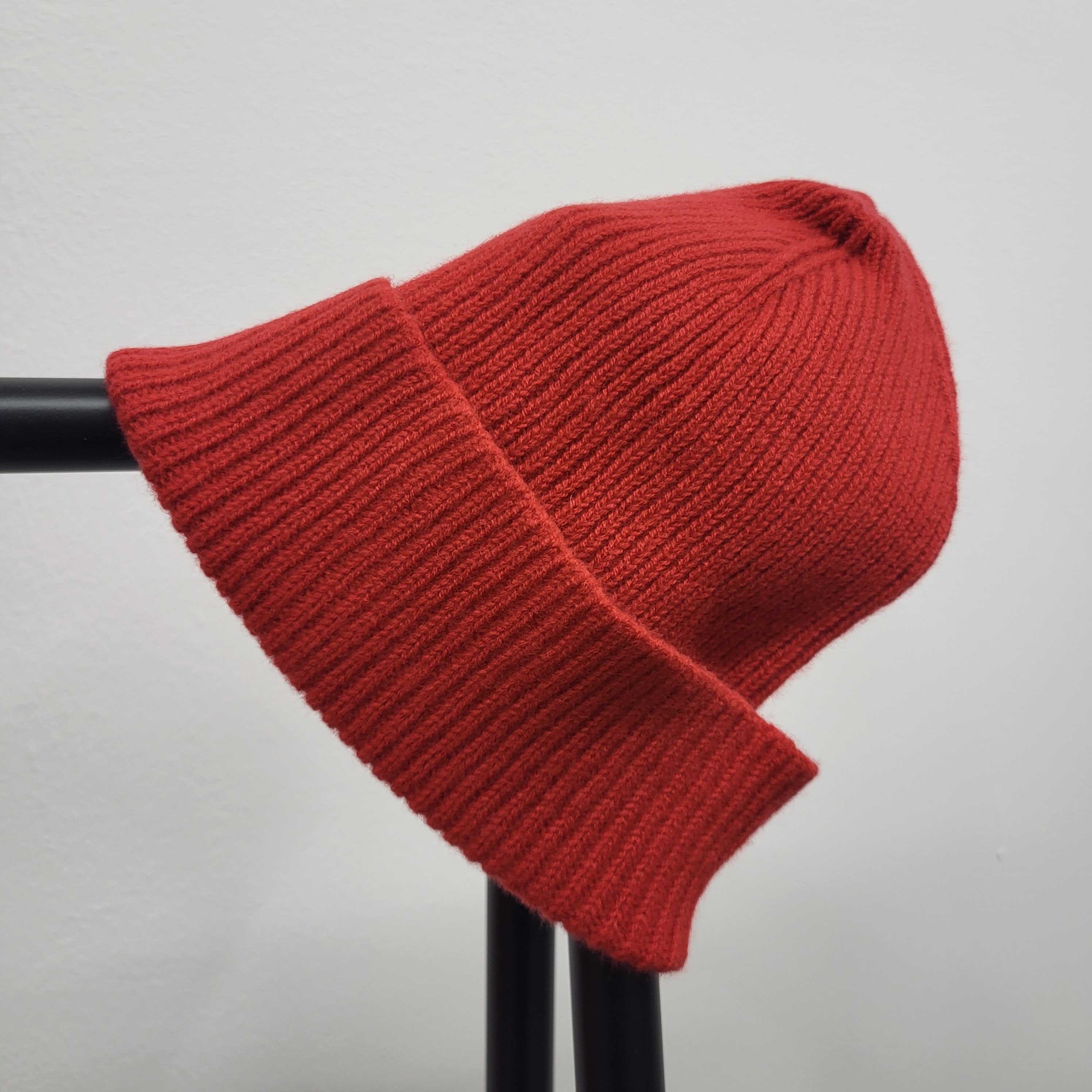 Rote, gestrickte Mütze liegt auf einem schwarzen Gestell vor einer weißen Wand.