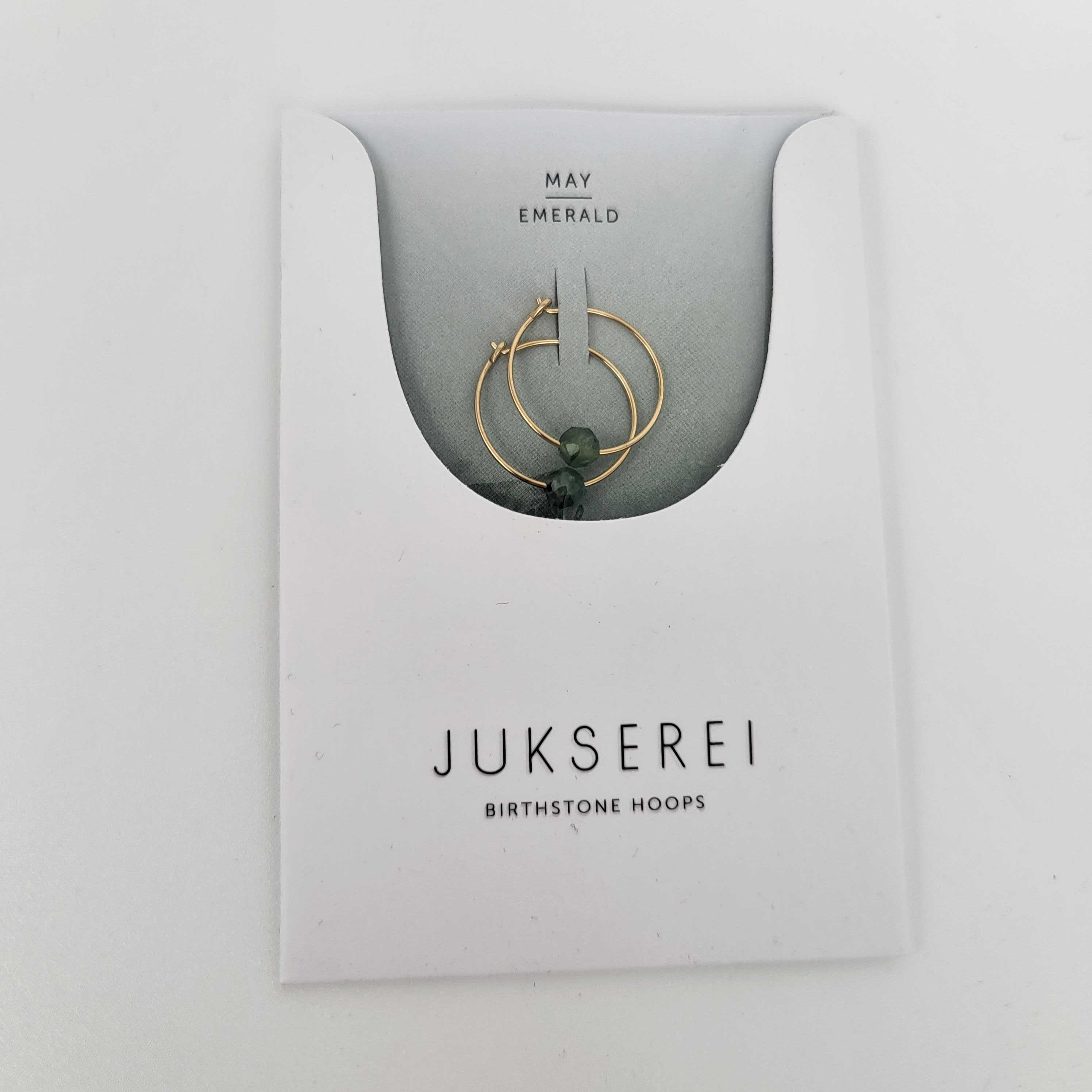 Goldene Ohrringe mit grünen Edelsteinen in einer JUKSEREI-Verpackung, beschriftet mit "May Emerald".