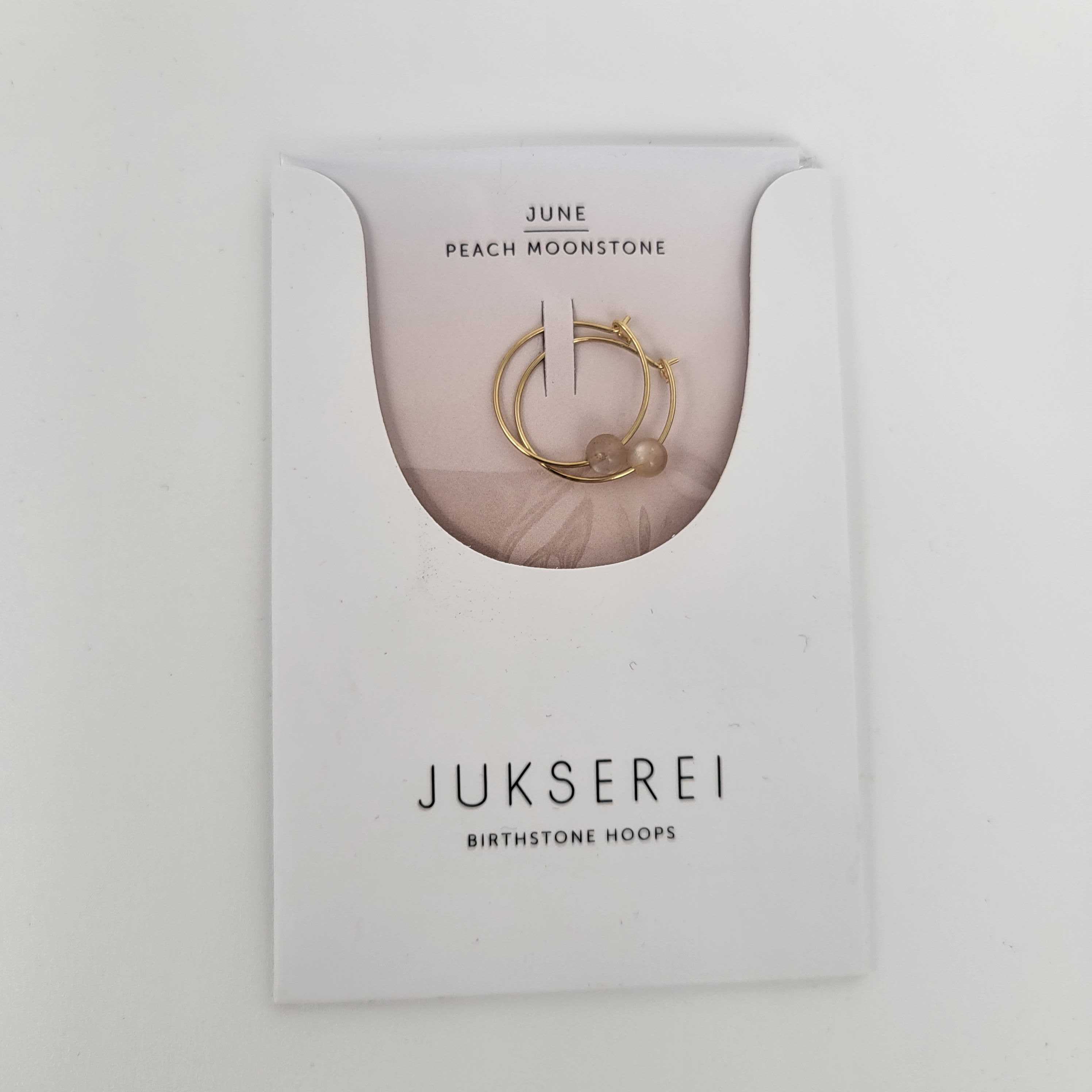Goldene Creolen mit Pfirsichmondstein auf einer Verpackung mit der Aufschrift "JUKSEREI Birthstone Hoops - June".