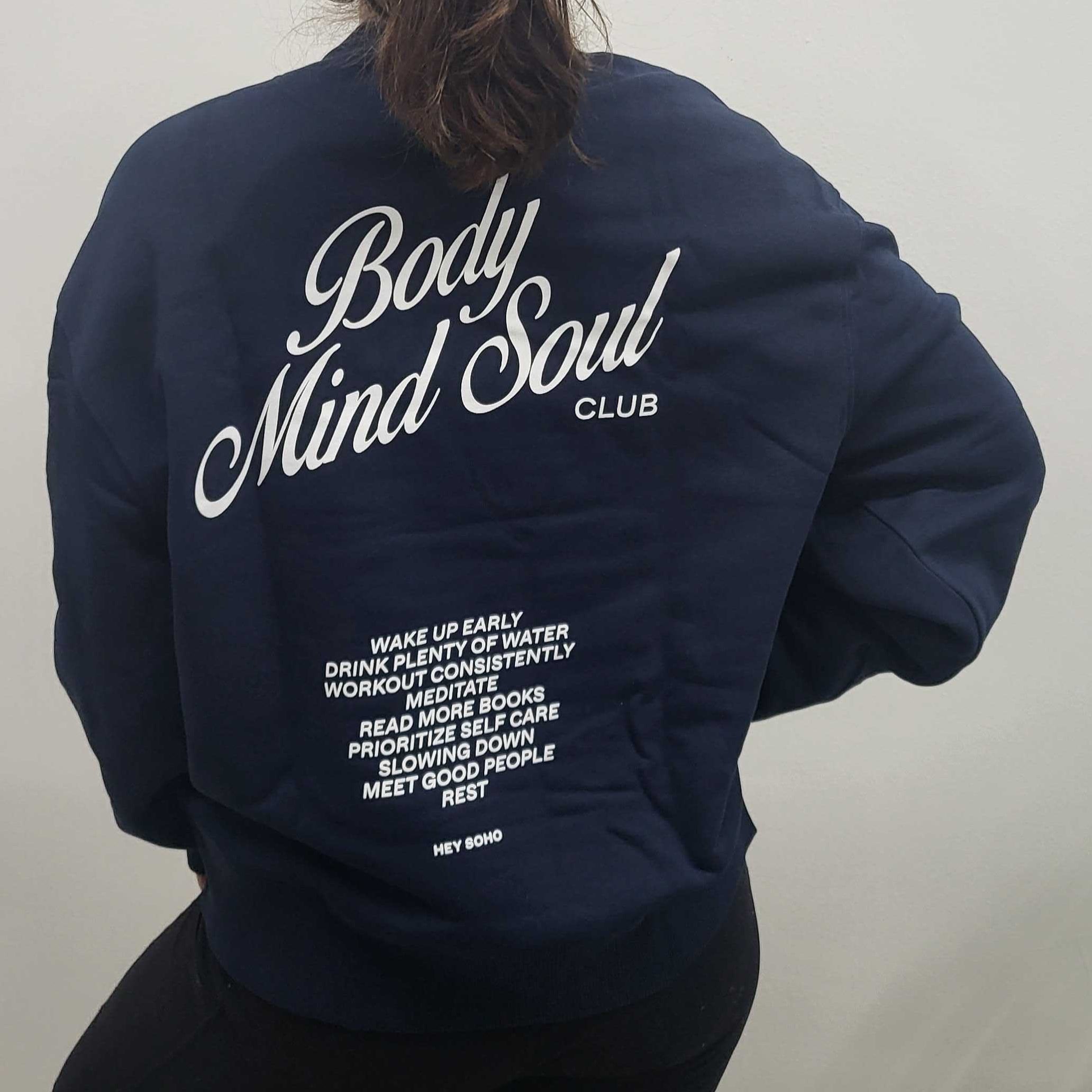 Frau mit blauem Sweatshirt, darauf steht "Body Mind Soul Club" und eine Liste von Selbstfürsorge-Tipps.