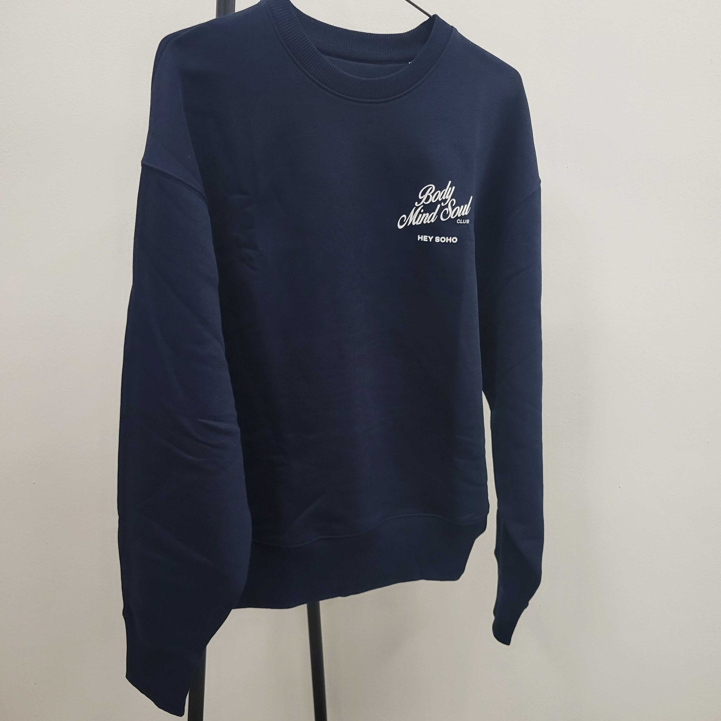 Dunkelblauer Pullover mit der Aufschrift "Body Mind Soul" und "HEY SOHO" in weißer Schrift, hängt an einem Kleiderständer.