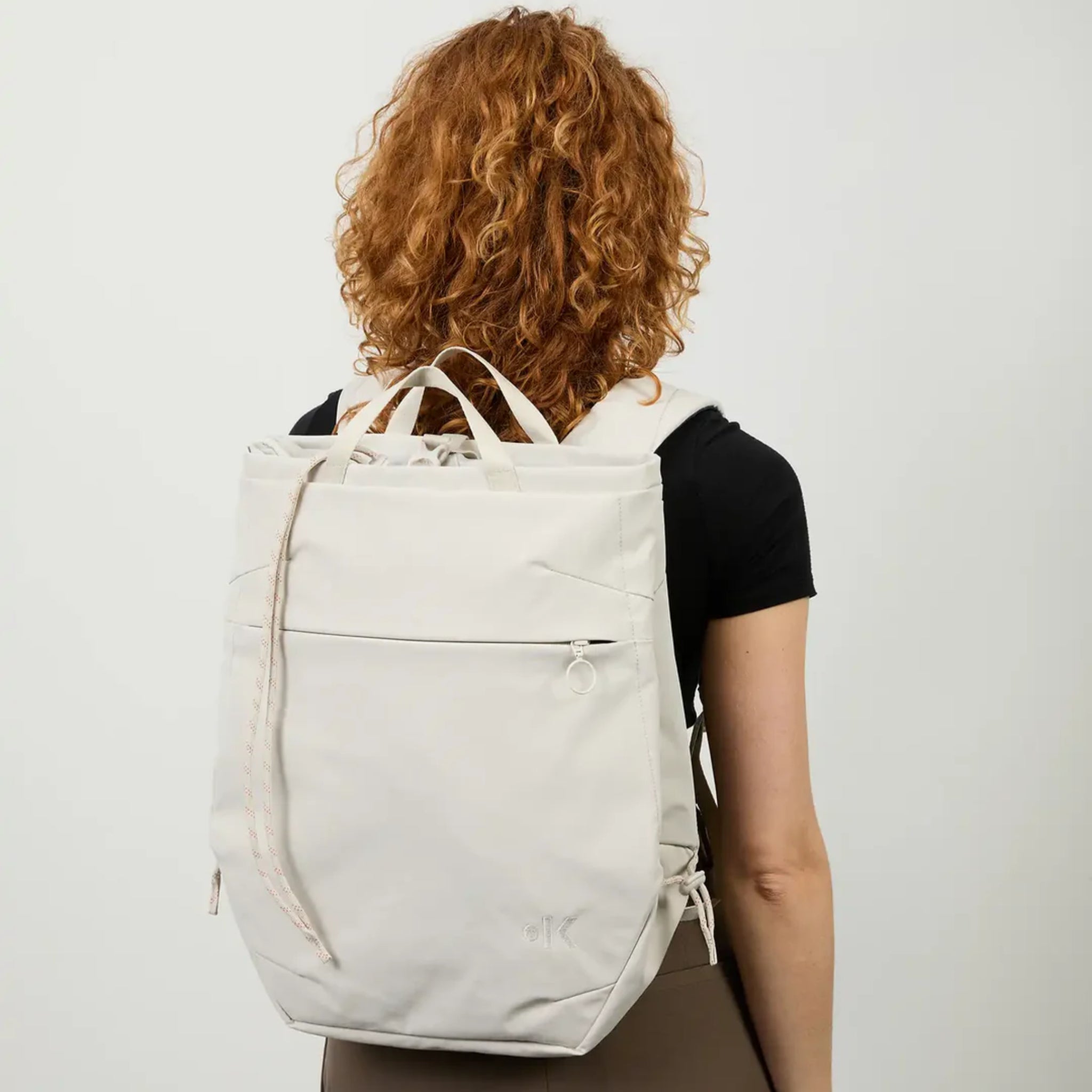 Eine Person mit roten Locken, die ein schwarzes Hemd trägt, wendet sich von der Kamera ab und trägt einen großen, cremefarbenen Rucksack mit kurzen Griffen und einer Reißverschlusstasche auf der Vorderseite.