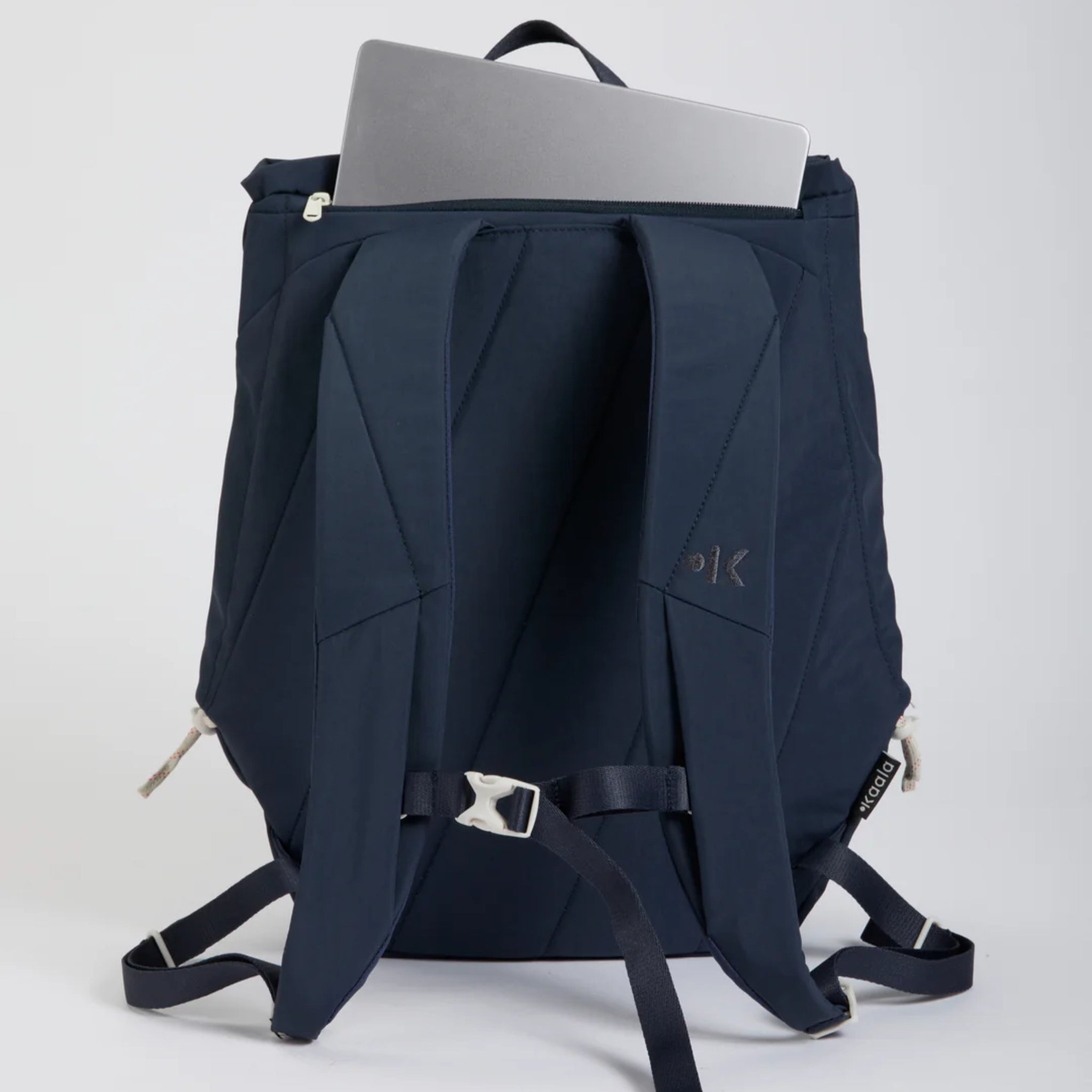 Ein marineblauer Rucksack mit gepolsterten Schultergurten sitzt aufrecht, oben ohne Reißverschluss, und im Inneren ist ein silberner Laptop teilweise sichtbar. Der Hintergrund ist einfarbig und hell.